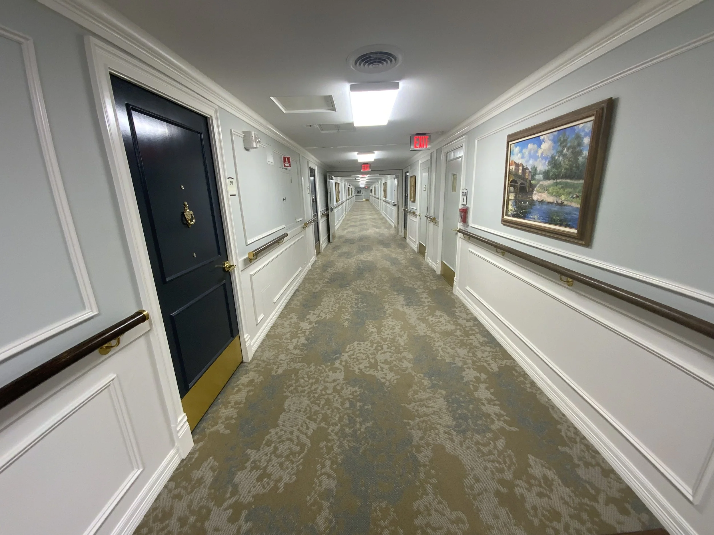A long hotel corridor with white wainscoting, brown handrails, patterned beige carpet, framed landscape paintings, and doors on either side. There are ceiling lights, a smoke detector, and visible exit signs.
