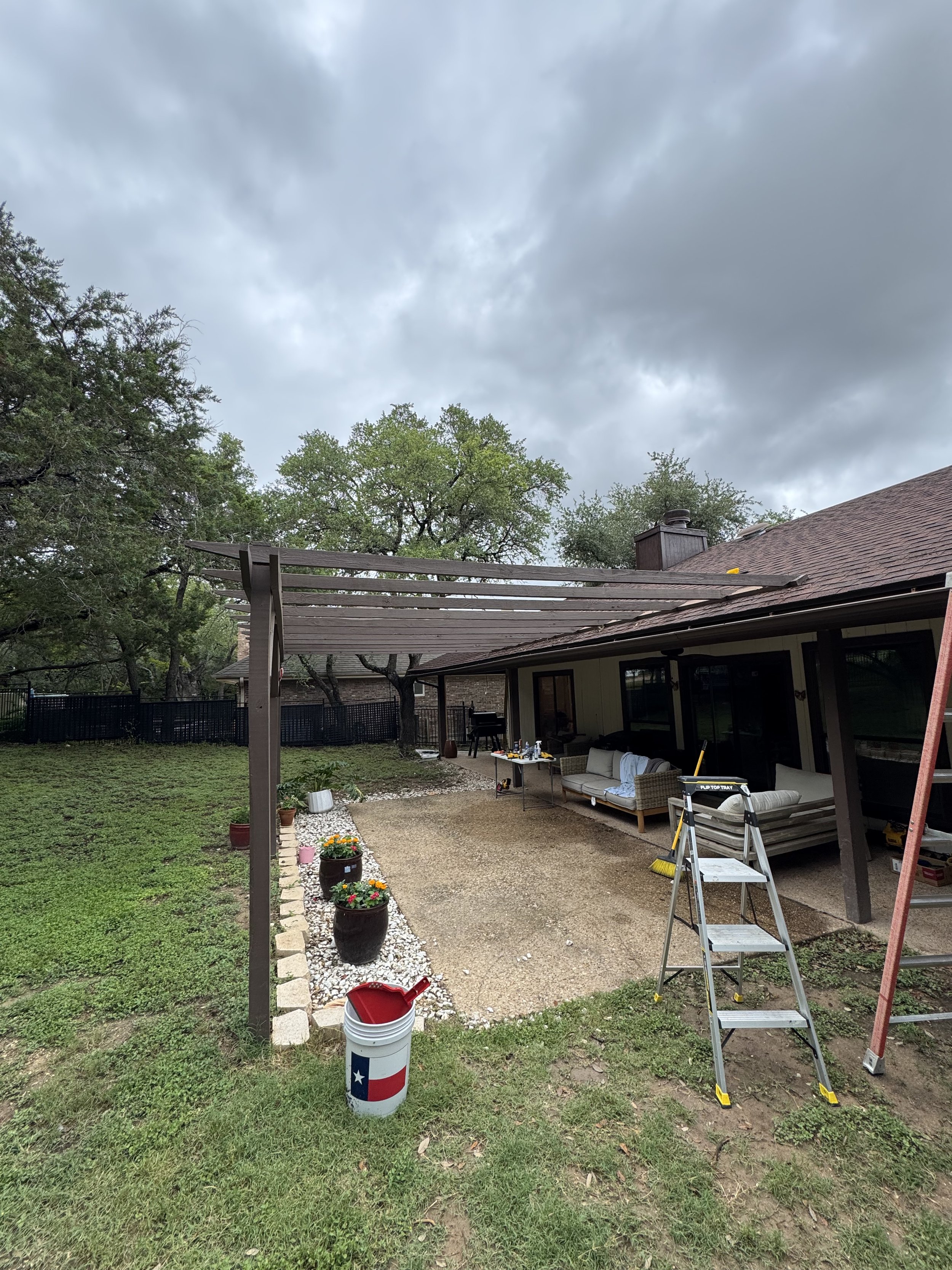 Backyard with a house, patio furniture, gardening tools, ladder, and potted plants, with cloudy sky overhead.