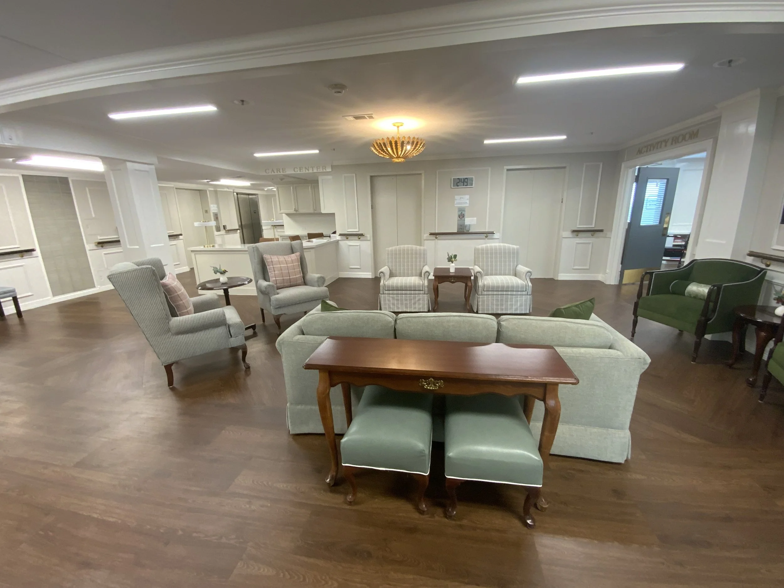 A waiting area with various chairs and small tables, a reception desk in the background, and a digital clock displaying 2:49 in a well-lit room with white walls and wood flooring.