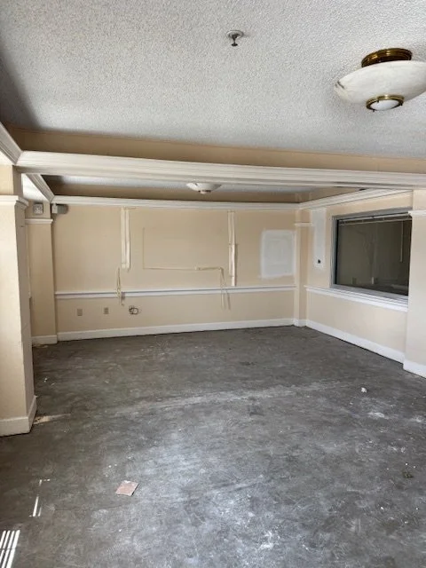 Empty room with beige walls, a large window, and ceiling light fixture, with a gray carpeted floor and some scattered debris.