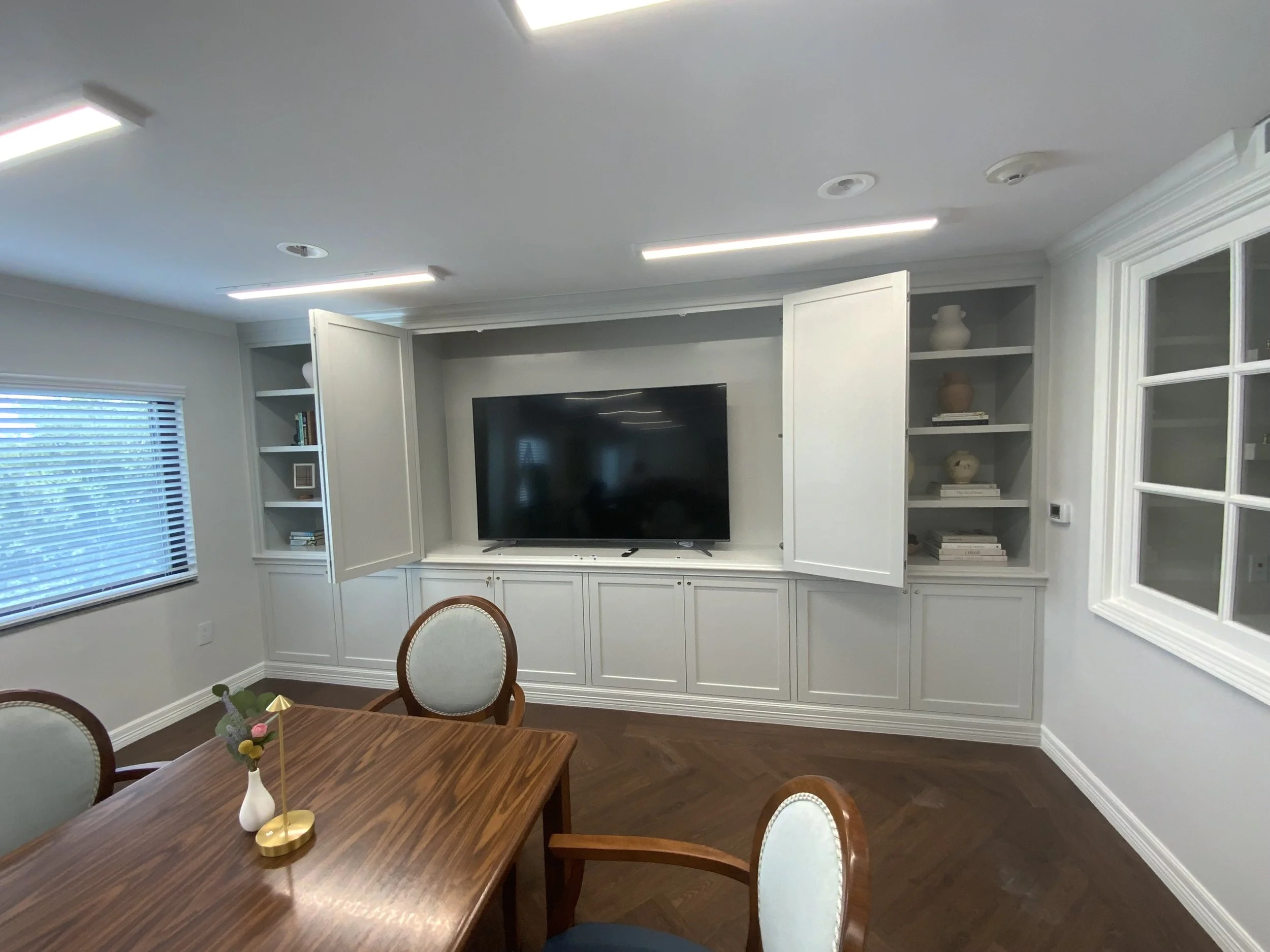 A living room with a white built-in entertainment center surrounding a large flat-screen TV. The room has white walls, a window with blinds, and dark hardwood flooring. A wooden dining table with chairs is in the foreground, and a small flower arrang
