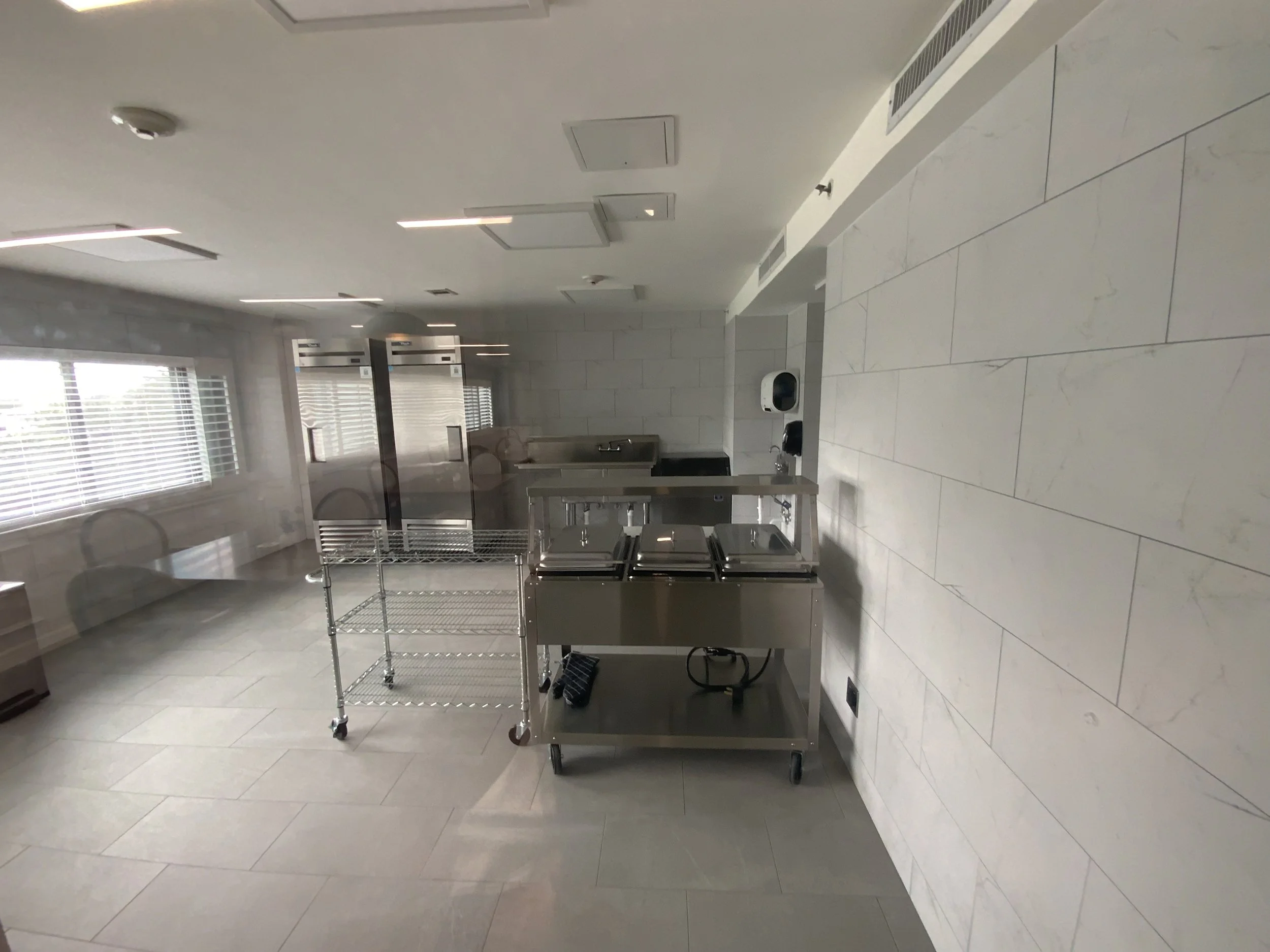 Empty room with white tiled floor and white walls, metal carts, a sink, and windows with blinds.