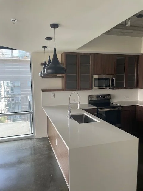 Modern kitchen with white island, dark wood cabinets, black pendant lights, stainless steel microwave, and oven, next to a large window with a sliding door.
