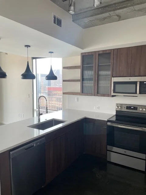 Modern kitchen with dark wood cabinets, white countertops, a microwave, oven, and a stainless steel sink, illuminated by hanging pendant lights.