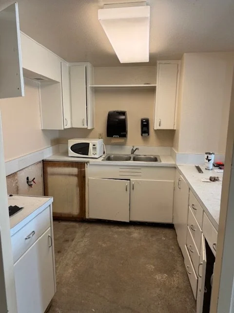 Small kitchen with white cabinets, a microwave, soap dispenser, and a sink. The floor is unfinished and some cabinet doors are missing or damaged.