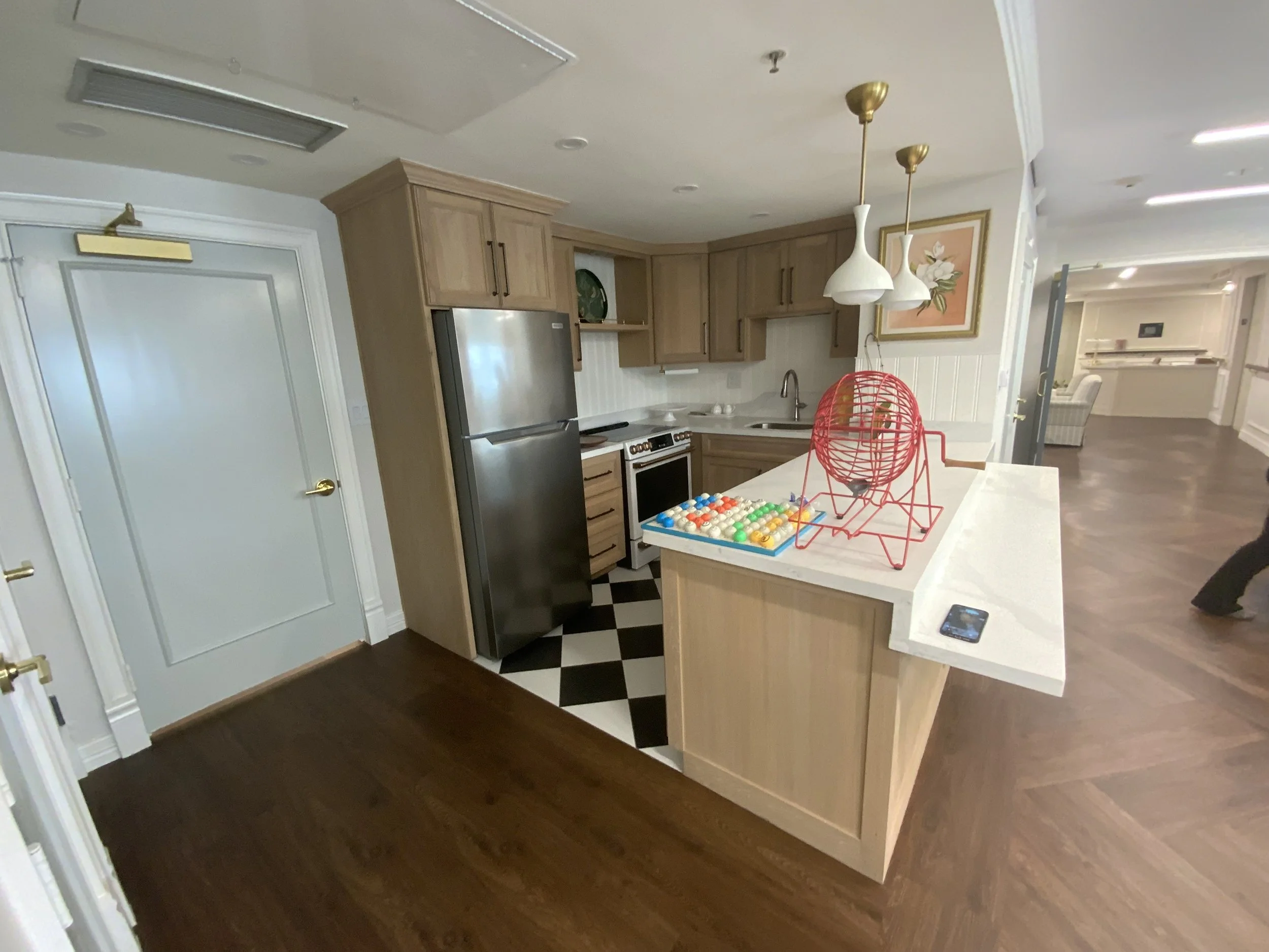 View of a modern kitchen with light wood cabinets, stainless steel refrigerator, black and white checkered floor, white countertops, two white pendant lights, a framed floral artwork, and a white island with various items including a red wire fruit b