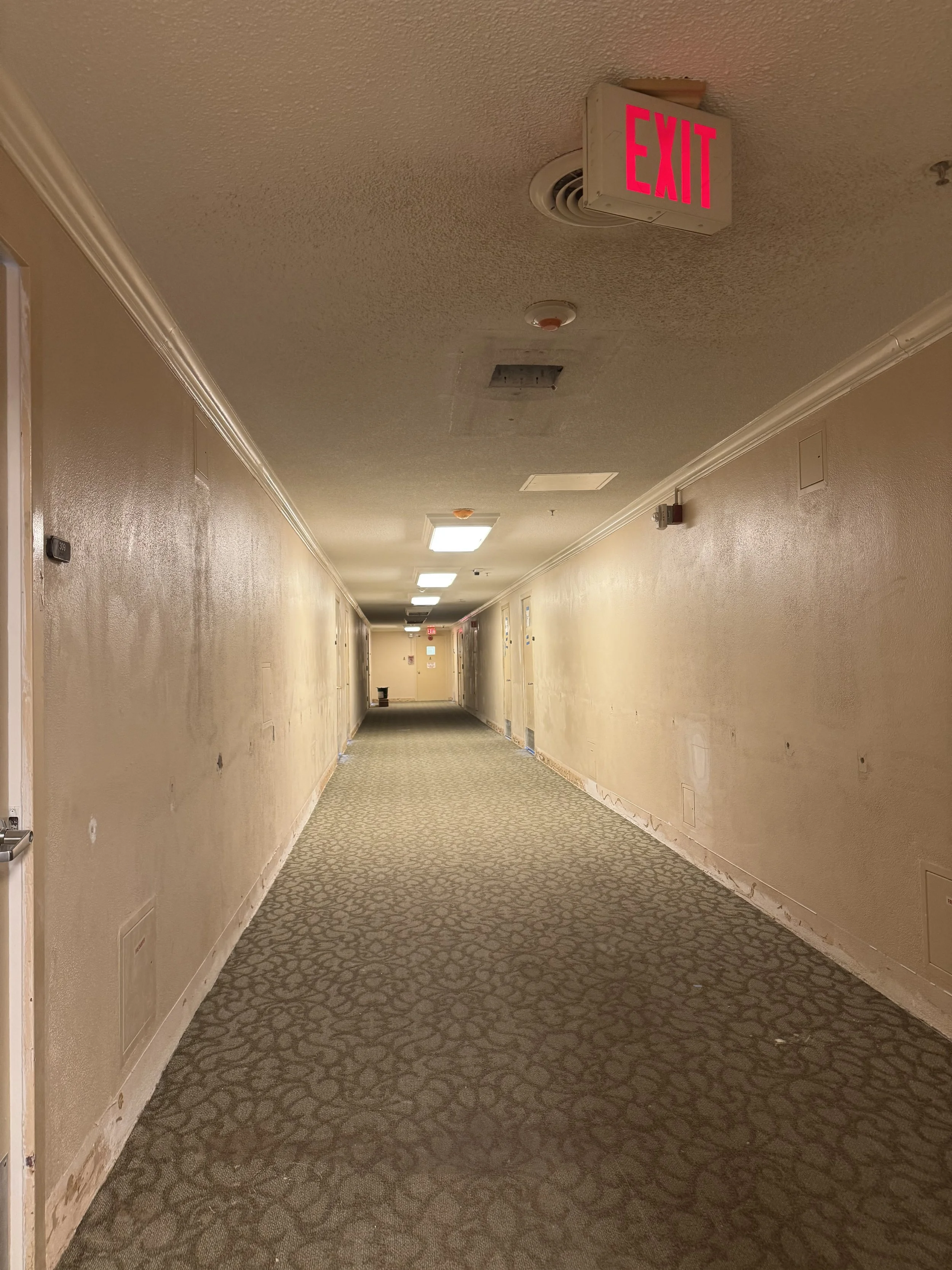 Empty hotel corridor with beige walls, patterned carpeting, and ceiling lights, showing signs of renovation.