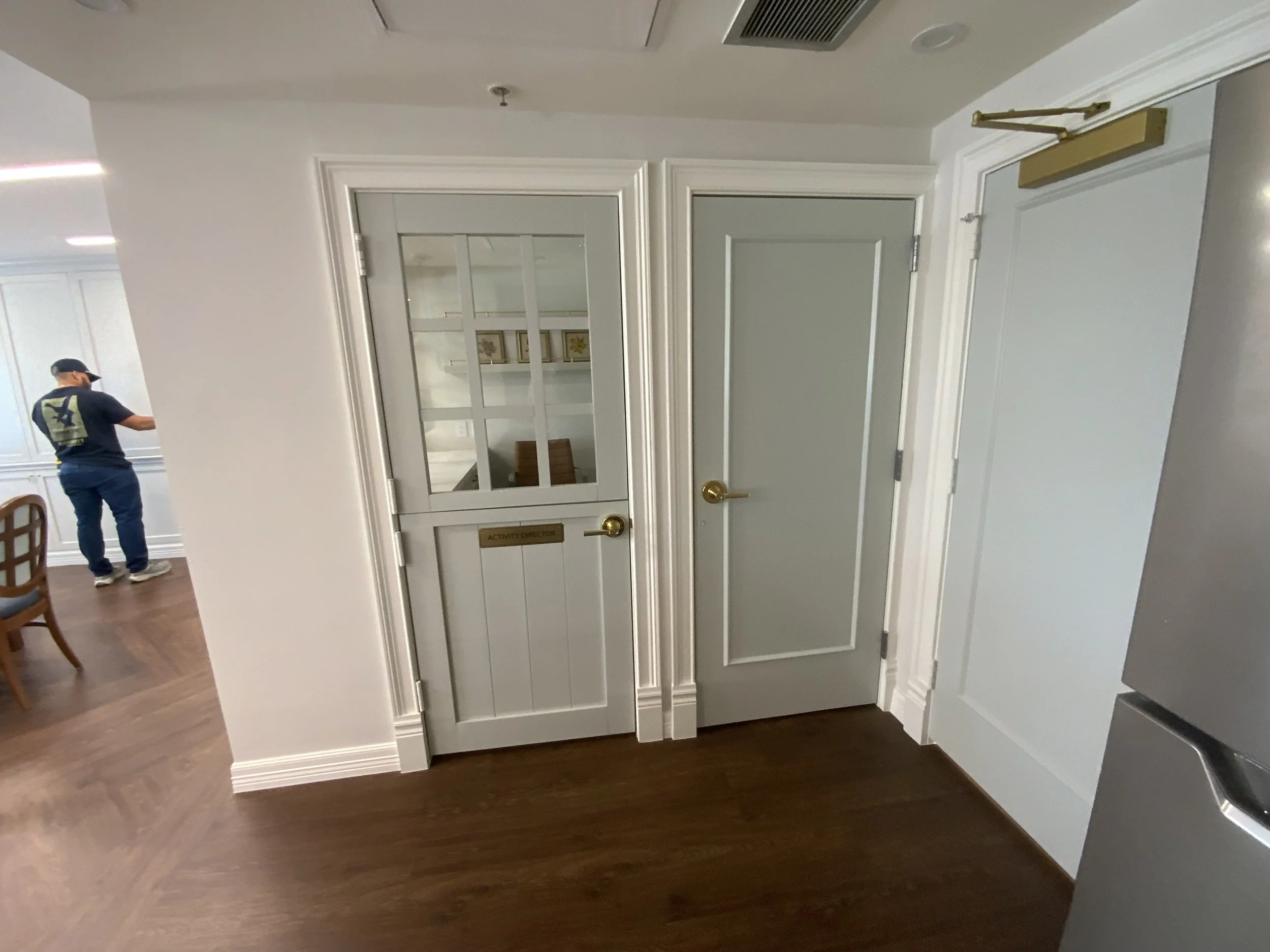 Interior of a room with a gray door marked 'Activity Director' and a glass window, hardwood floors, and part of a white wall with crown molding. A person in dark clothing and a cap is walking in the background.