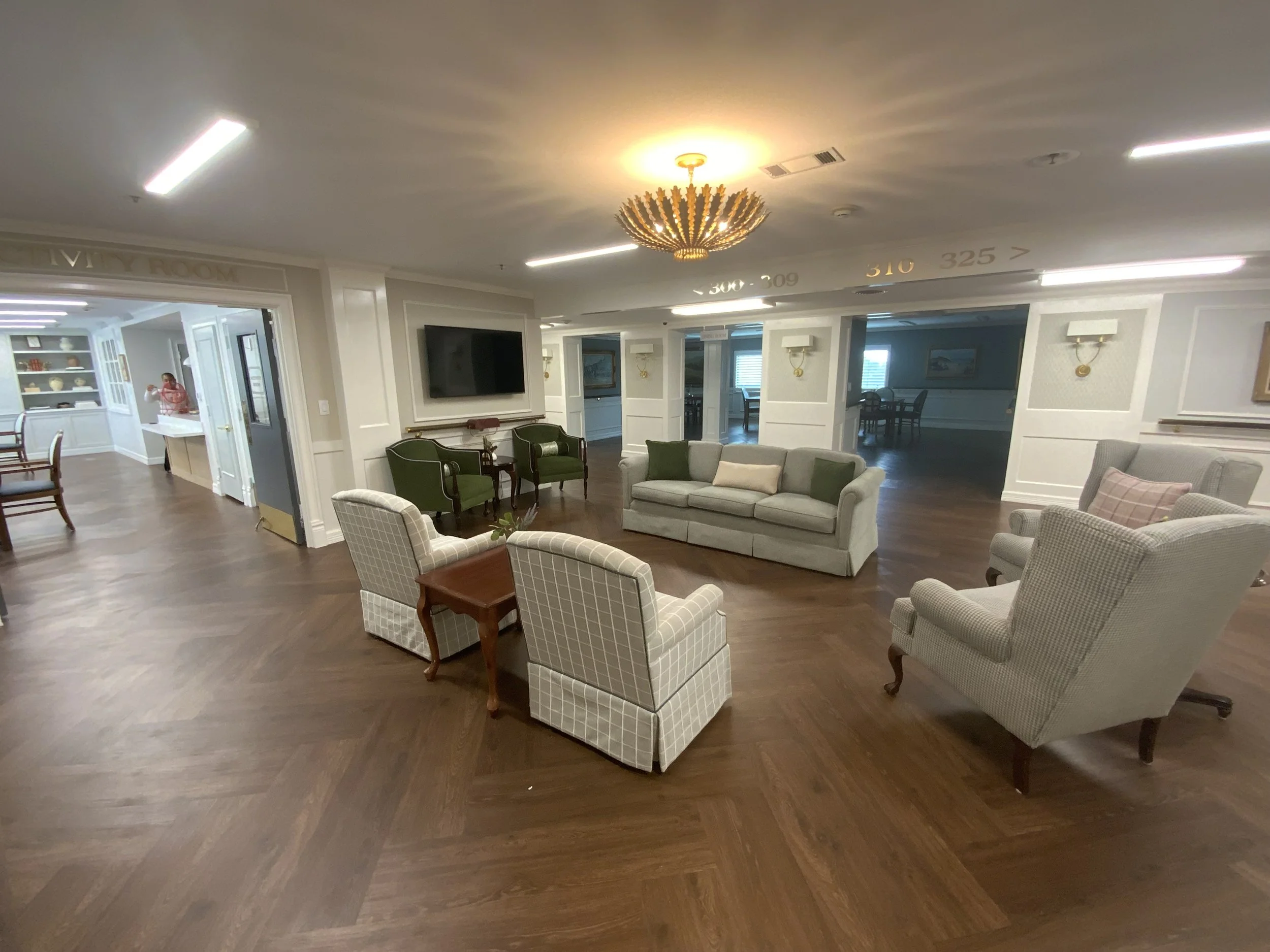 A living room with a chandelier, several armchairs, a sofa, a wall-mounted television, and a dining area visible through an open door, with hardwood floors and white walls.
