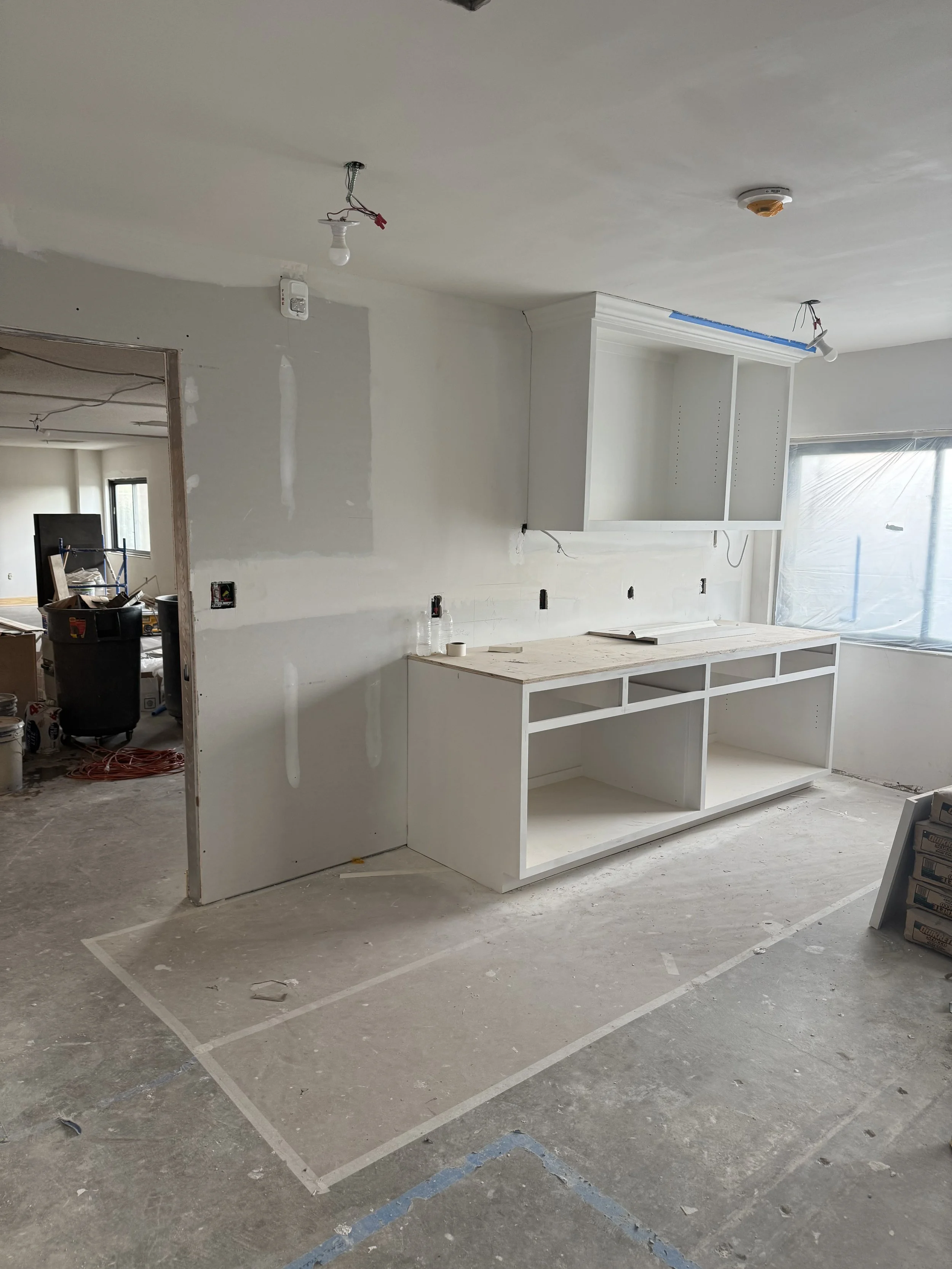 A room under construction with unfinished walls and ceiling, some drywall work, a white built-in cabinet with open shelves, and tools and materials scattered on the floor.