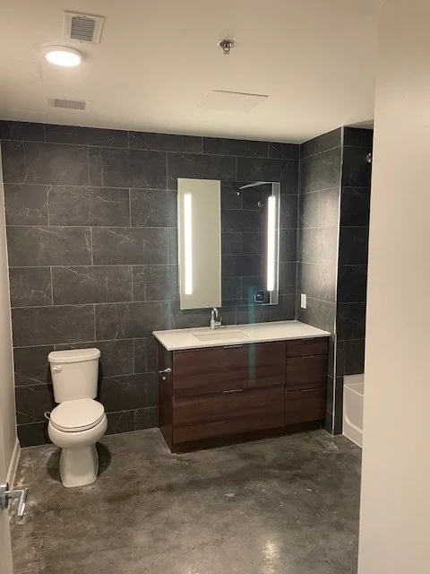 Modern bathroom with dark gray tiled walls, a white toilet, a wooden vanity with a sink, a large mirror with LED lights, and a bathtub on the right.