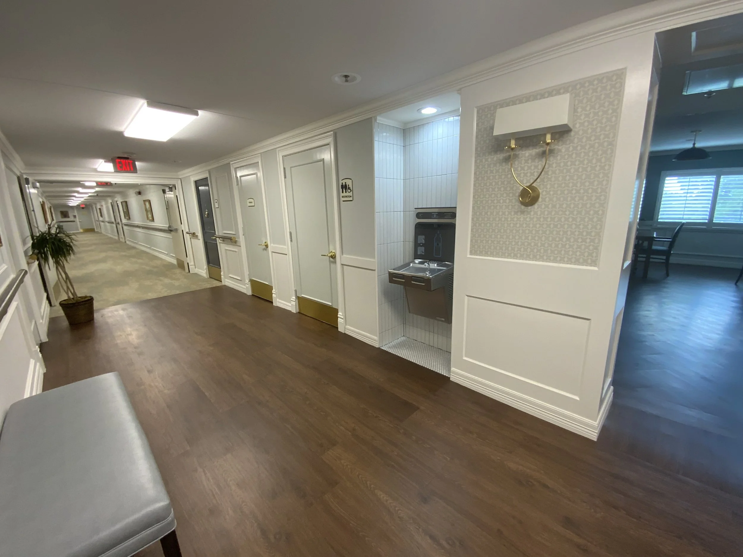 Empty hallway in a building with white walls, wooden floors, and decorated with paintings. There is a potted plant on the left side and signs indicating restrooms. On the right side, there is a water fountain. The hallway has a lit ceiling and an exi