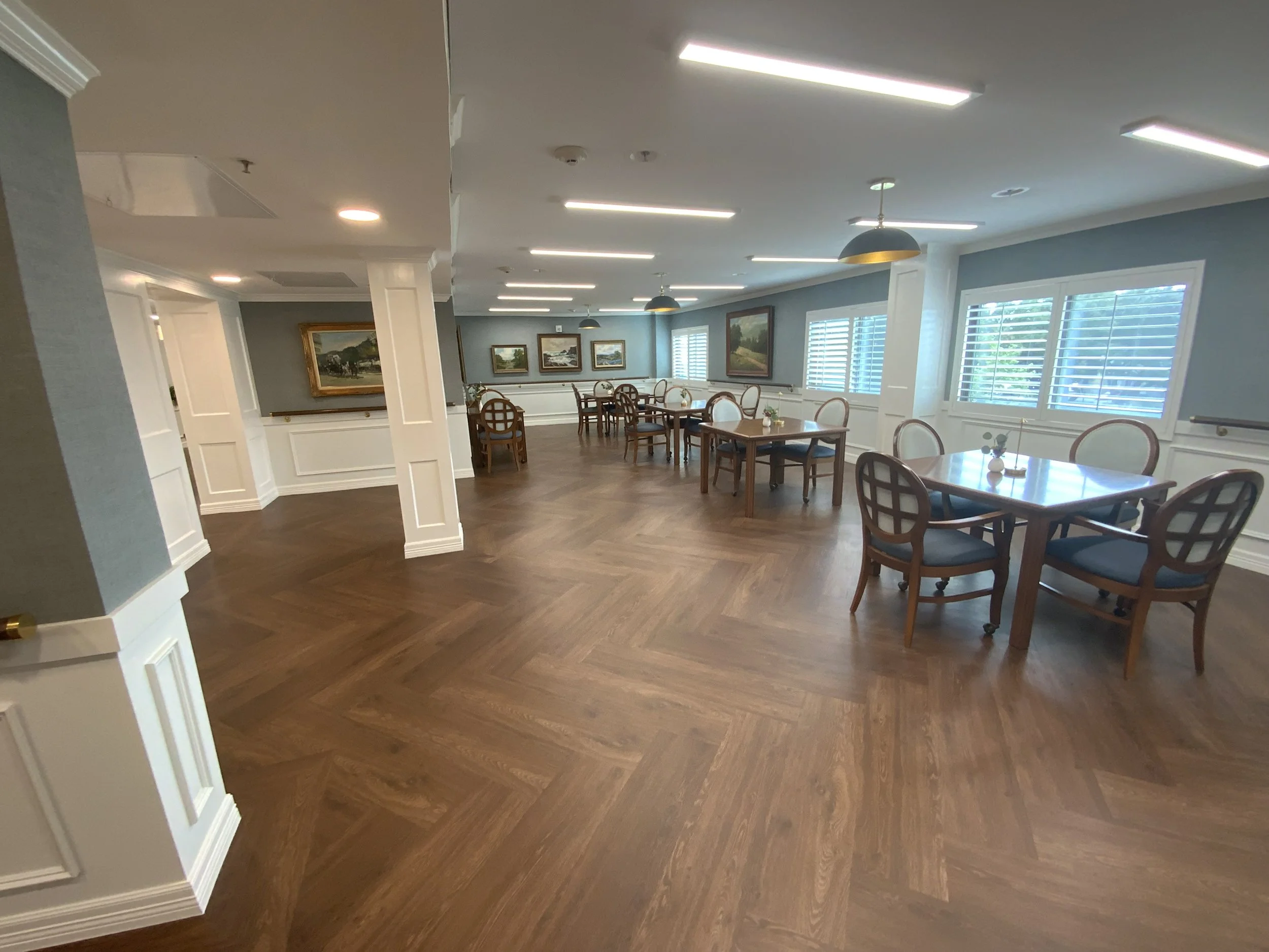 A bright, spacious room with wooden flooring, white-paneled walls, and multiple windows with white shutters. Several tables and chairs are neatly arranged around the room, which is decorated with framed landscape paintings on the walls. The ceiling h
