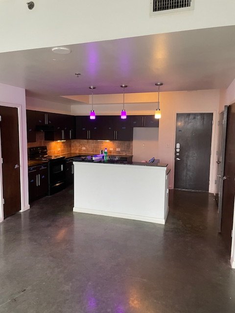 Empty kitchen and living area with a white counter, dark cabinets, pendant lights with colored bulbs, and a black door.