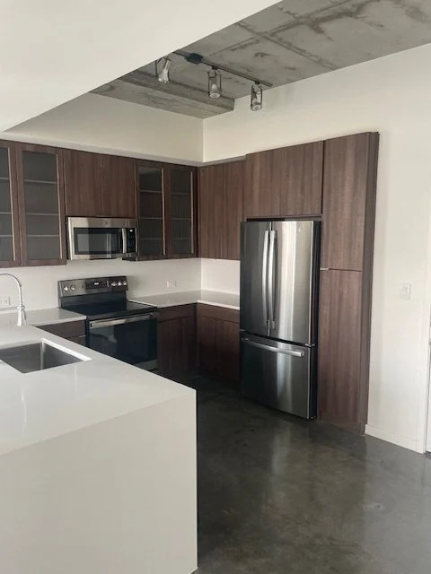 Modern kitchen with wooden cabinets, stainless steel refrigerator, oven, microwave, and white countertop with sink.