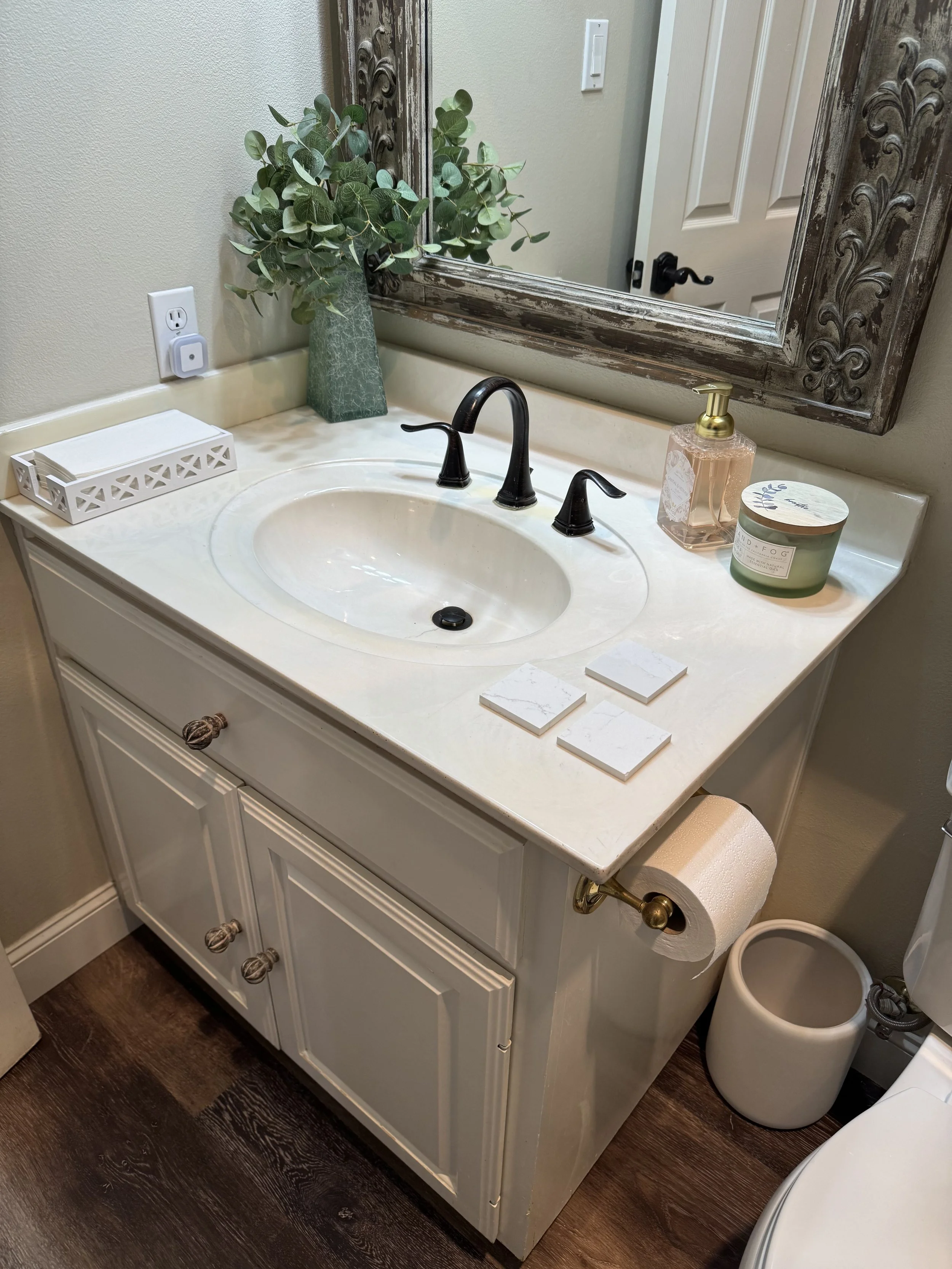 Bathroom vanity with a sink, faucet, decorative mirror, potted plant, soap dispenser, candles, toilet paper, and trash can.