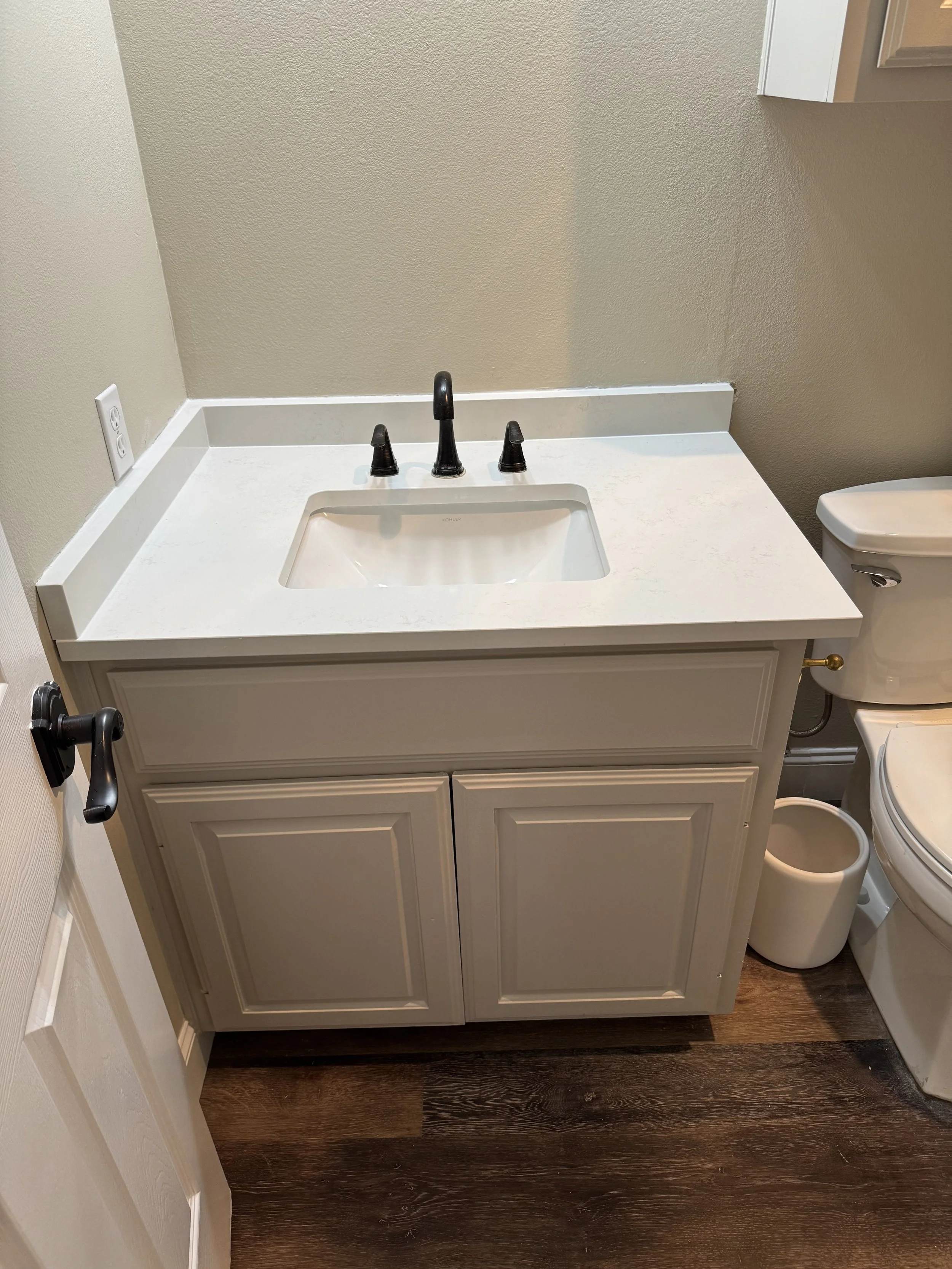 Bathroom vanity with a white countertop, black faucet, and two cabinet doors beneath. To the right, a toilet and small trash can are visible. The floor is dark wood.