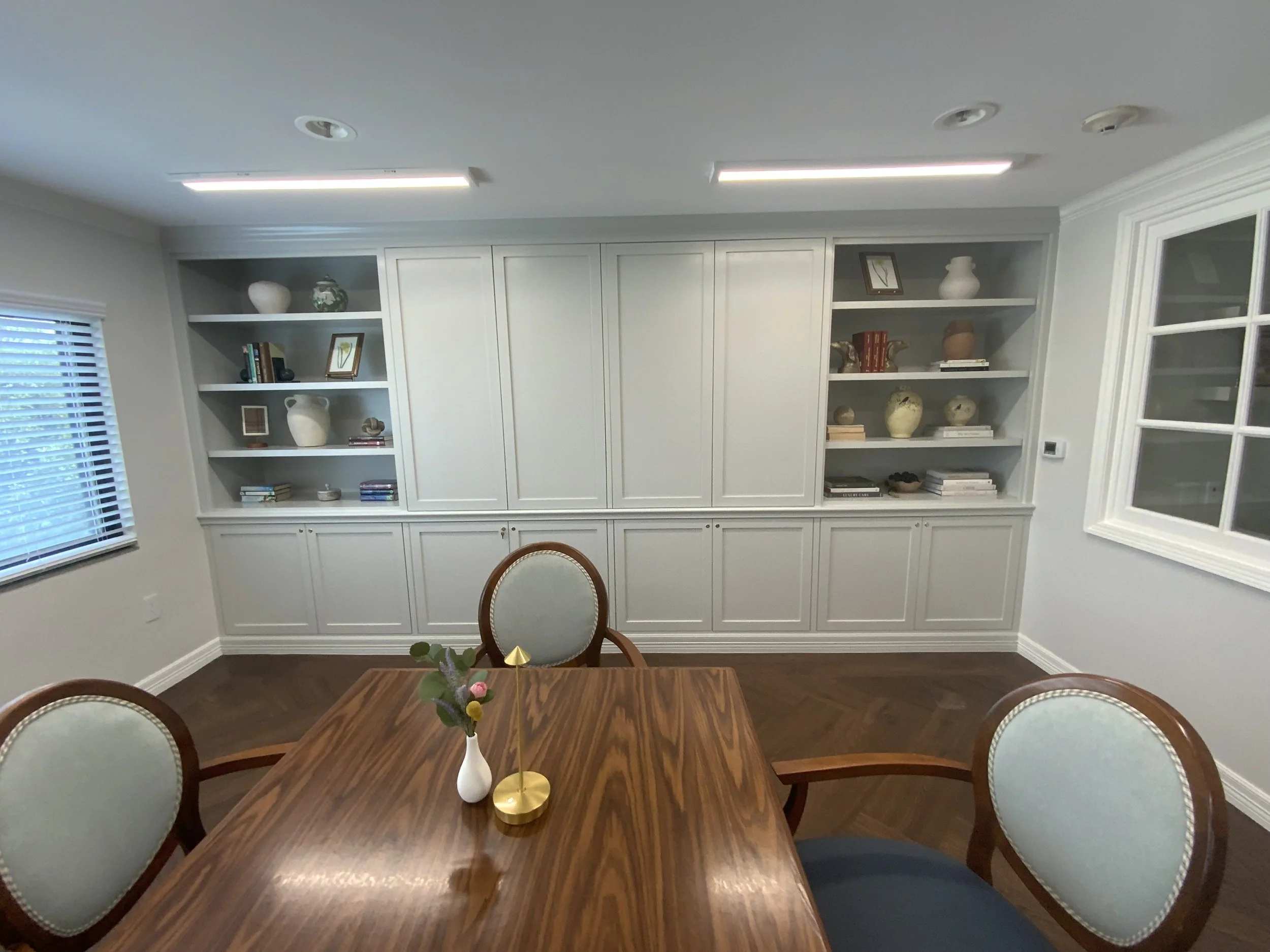 A dining room with a wooden table and four chairs, a cabinet with shelves filled with decorative items and books, a window with blinds, and a small flower arrangement on the table.
