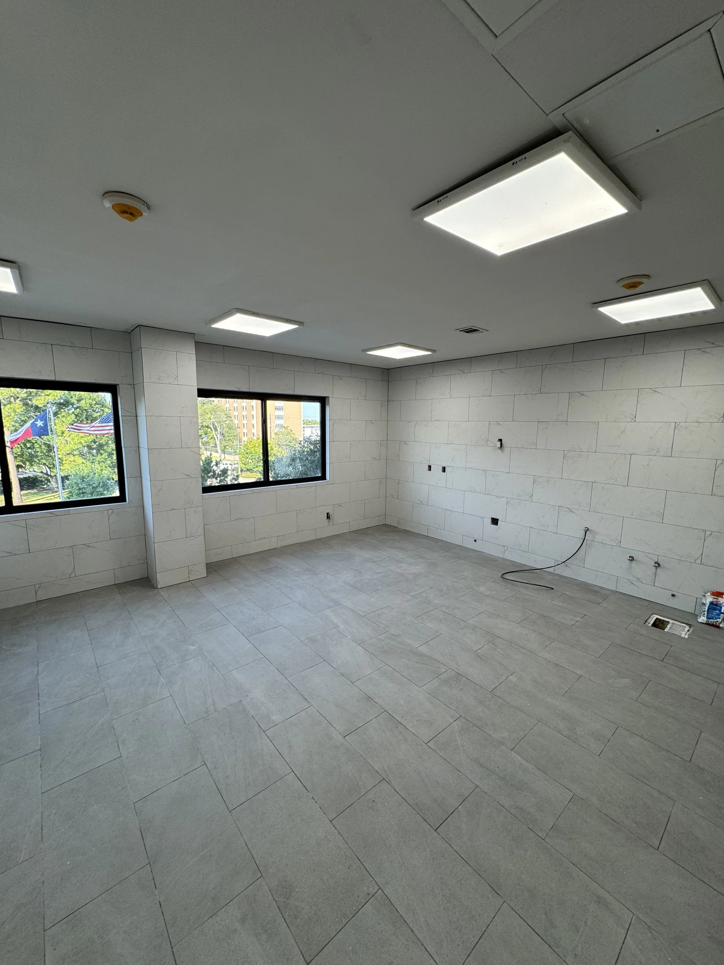 Empty room with tiled floor and white brick walls, windows showing trees and flags outside, ceiling lights, and some electrical wiring and outlets on the wall.
