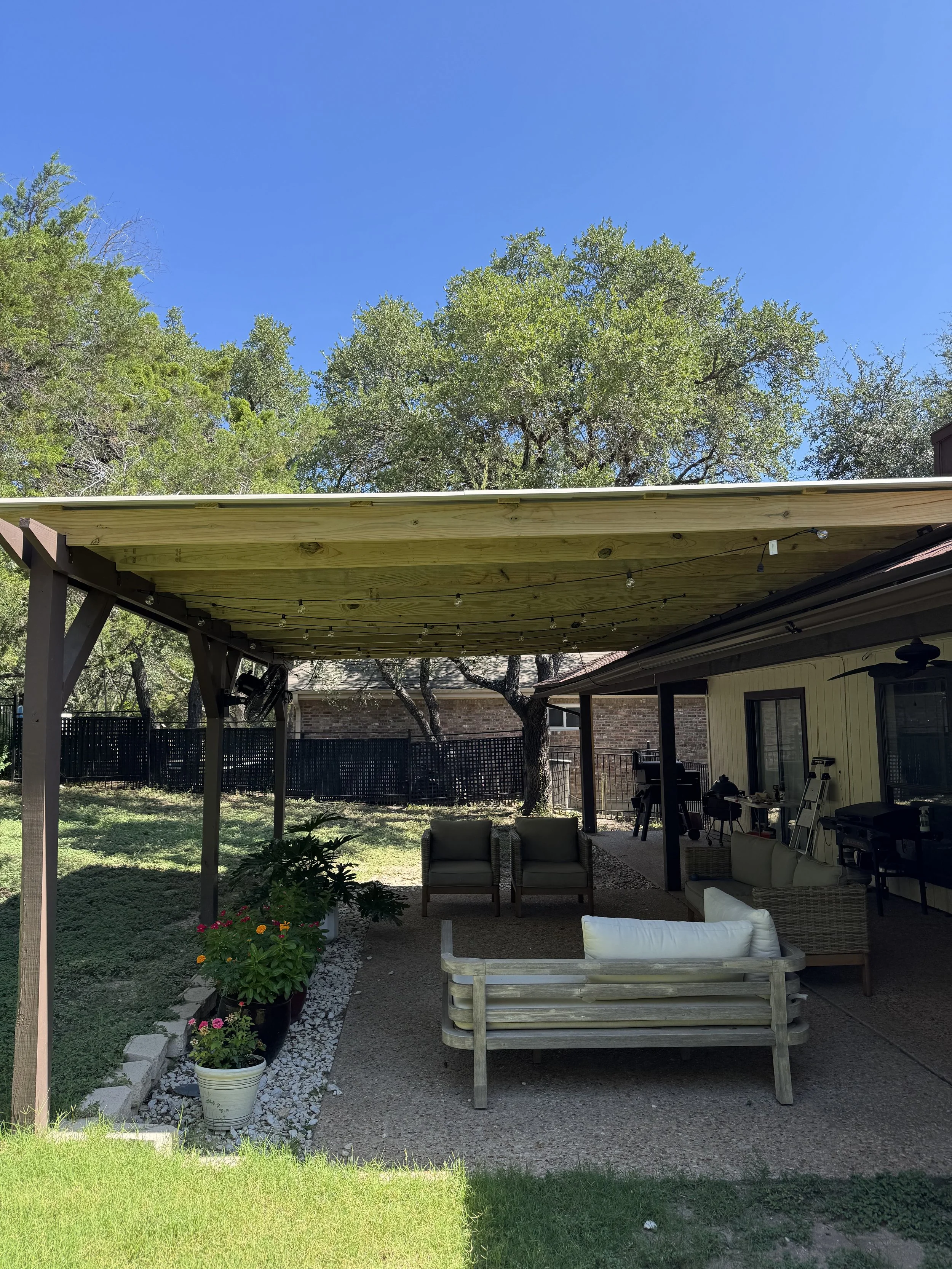 A backyard patio with outdoor furniture, potted plants, trees, and a clear blue sky overhead.