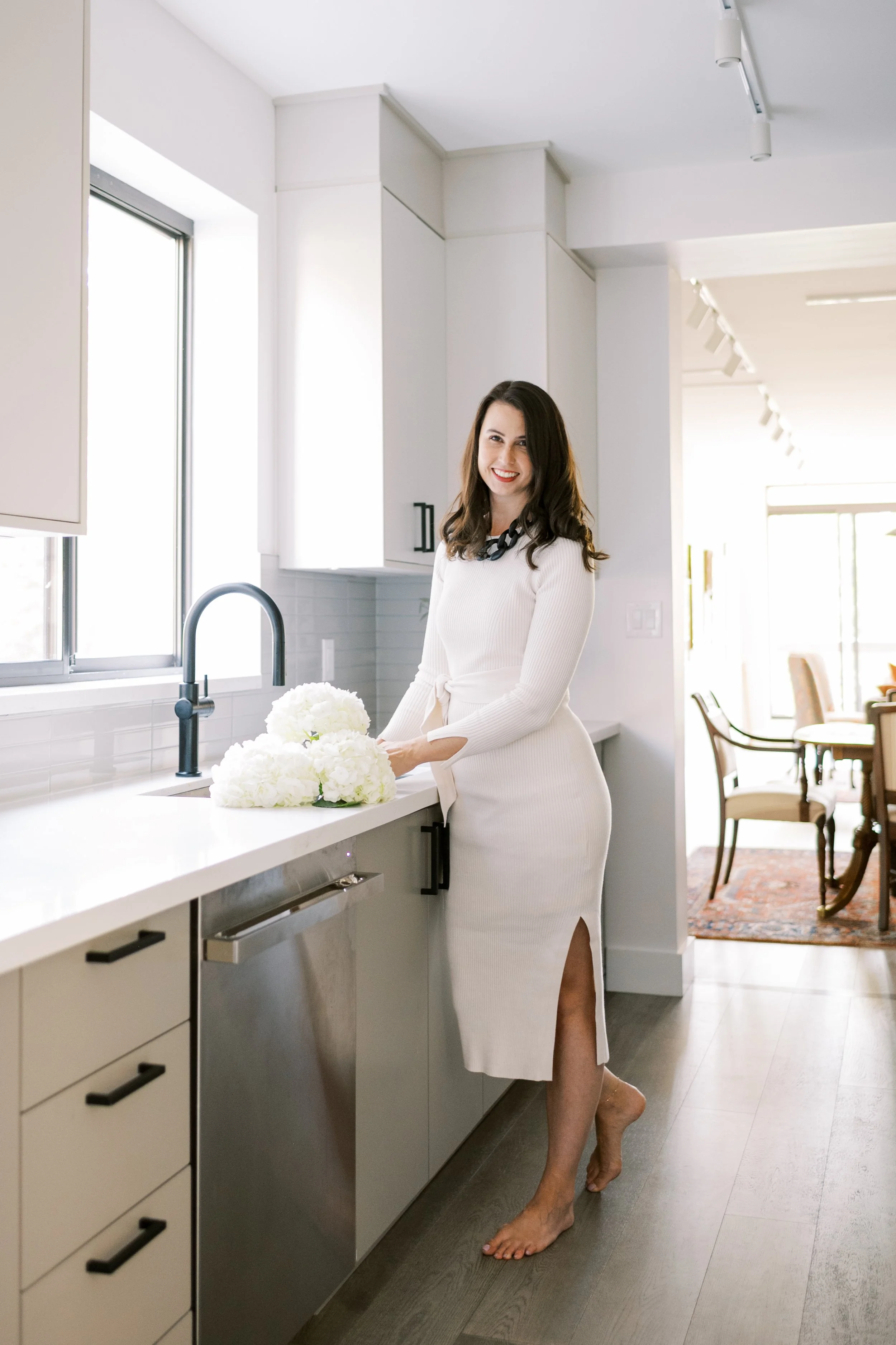 A woman with dark brown hair wearing a white, long-sleeve, fitted dress with a slit, is standing barefoot in a modern kitchen. She is smiling and washing white hydrangeas in a sink next to a window.