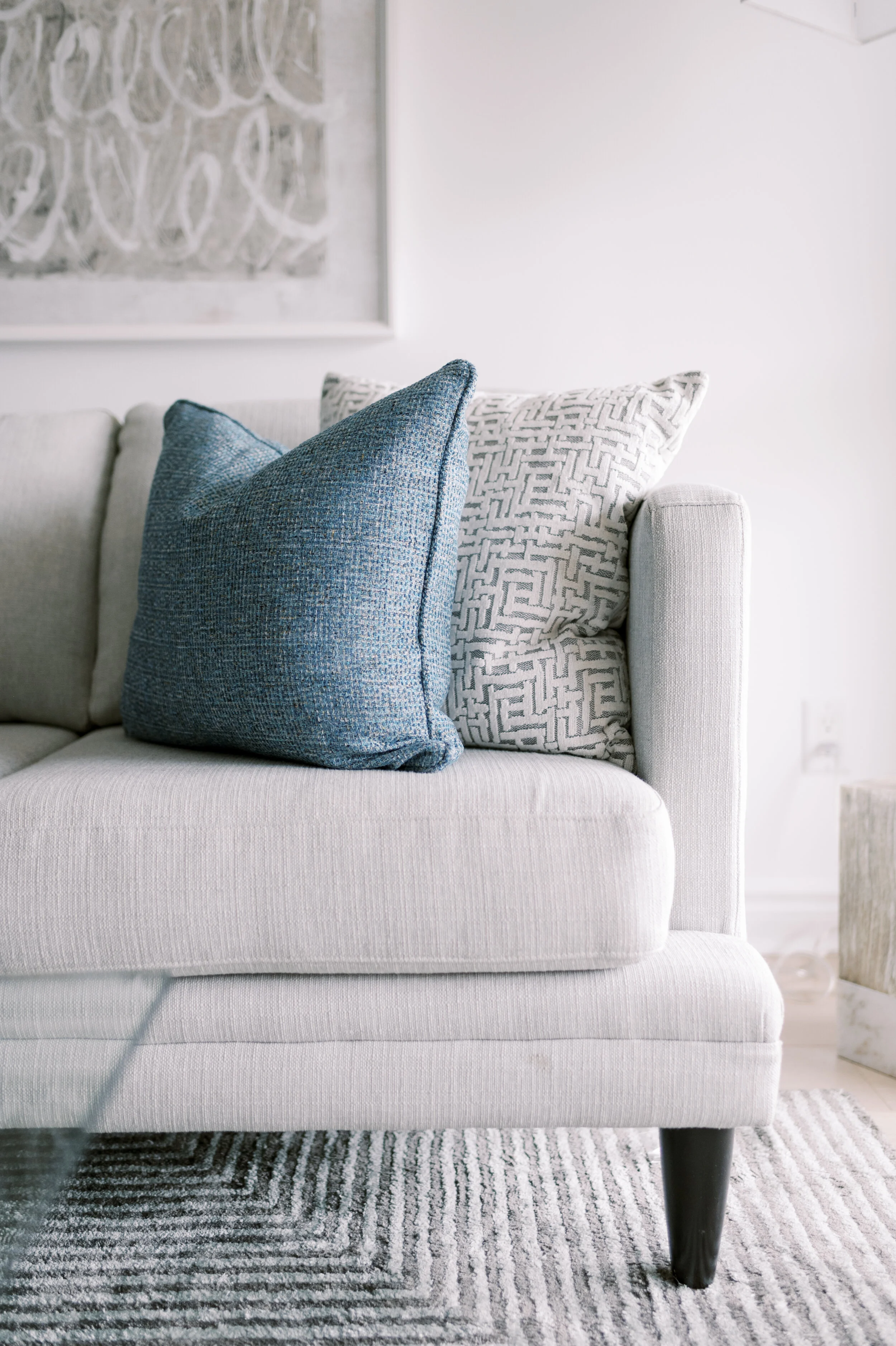 Close-up of a cream-colored sofa with two pillows, one dark blue and one white with a geometric pattern, in a modern living room with artwork on the wall and a gray patterned rug.