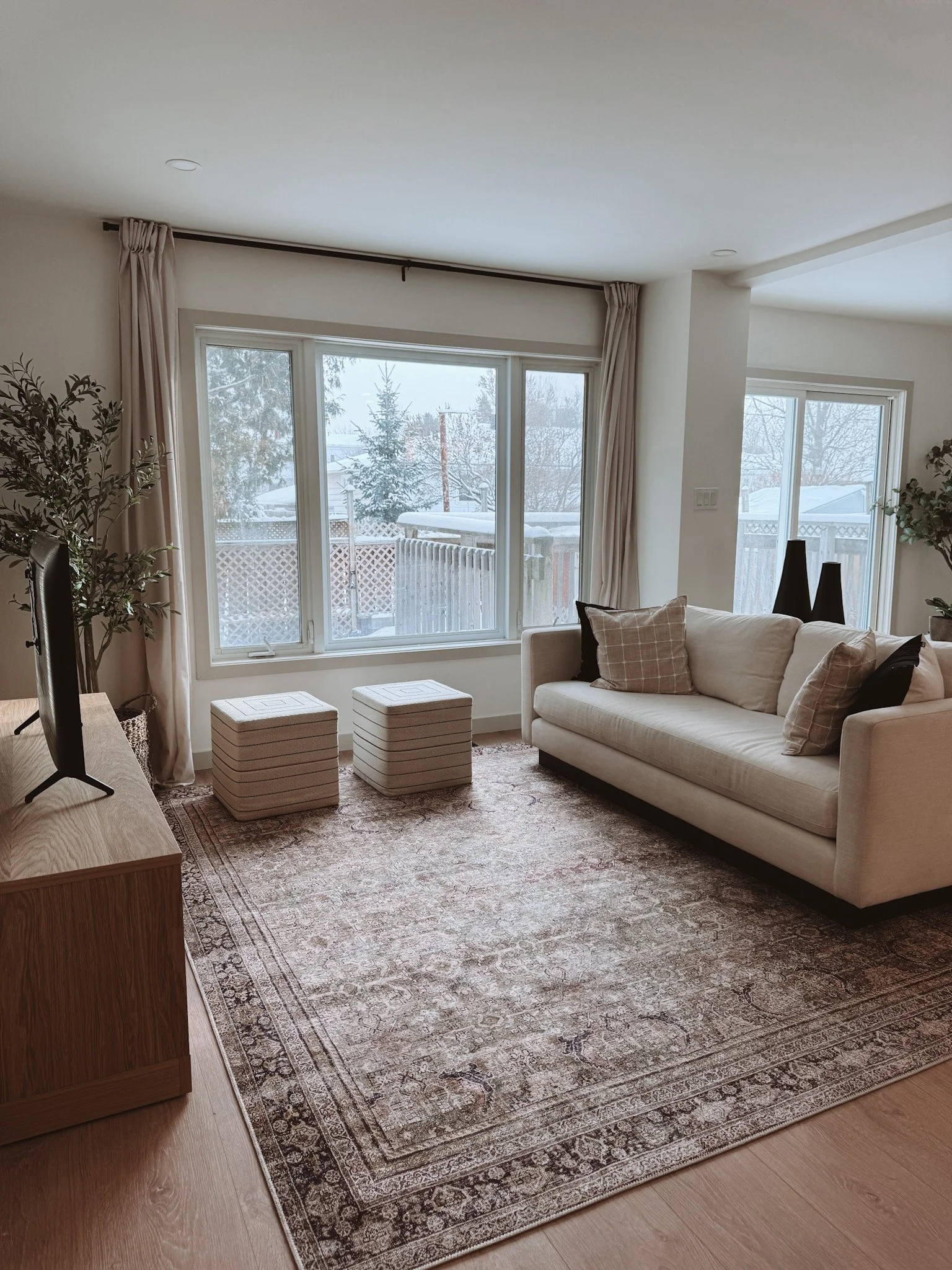 Living room with beige sofa, three throw pillows, window view of a snowy backyard, patterned area rug, two striped ottomans, and a wooden TV stand with a TV.