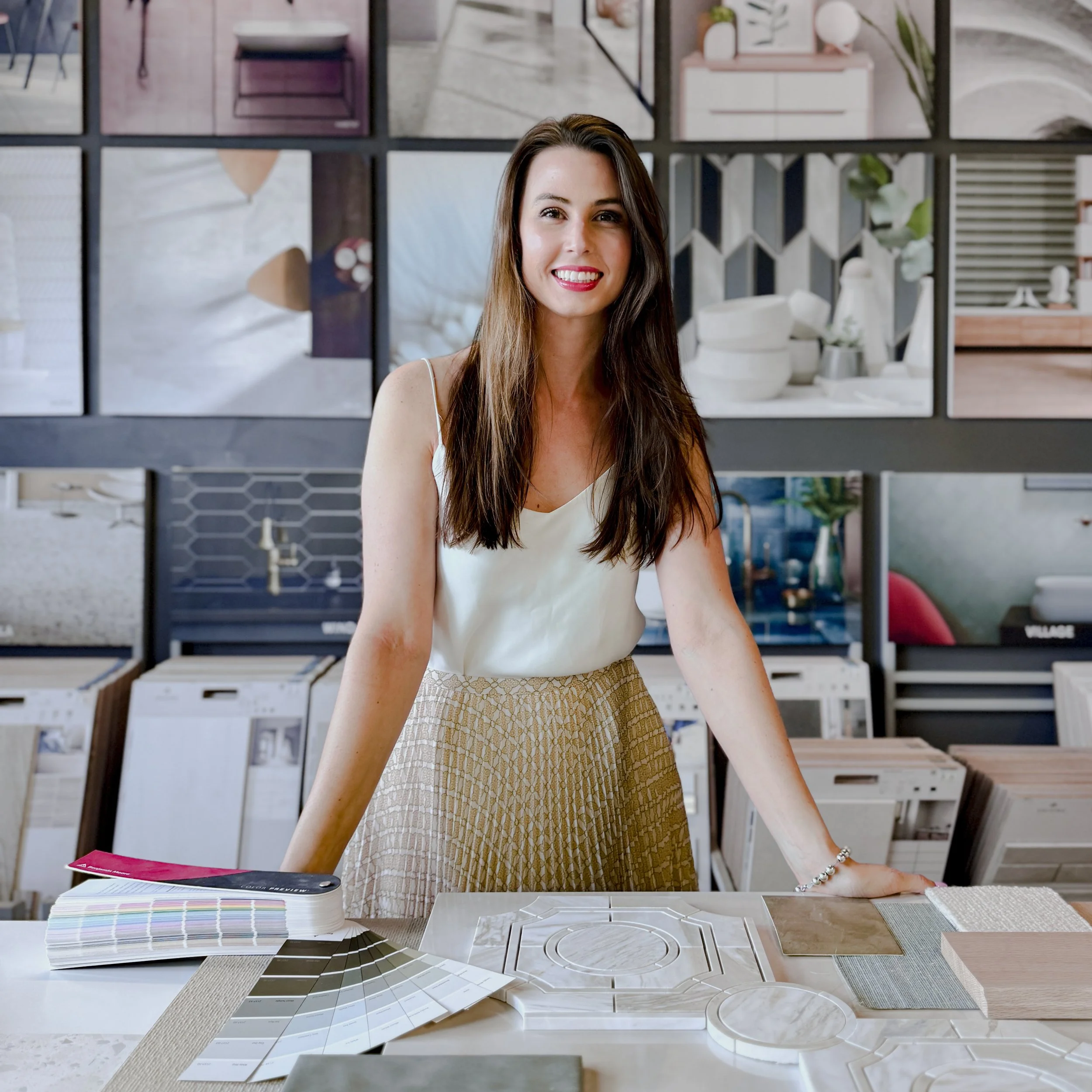 A woman smiling while standing behind a table with sample tiles, color swatches, and design materials in a store, with home decor and tile displays in the background.
