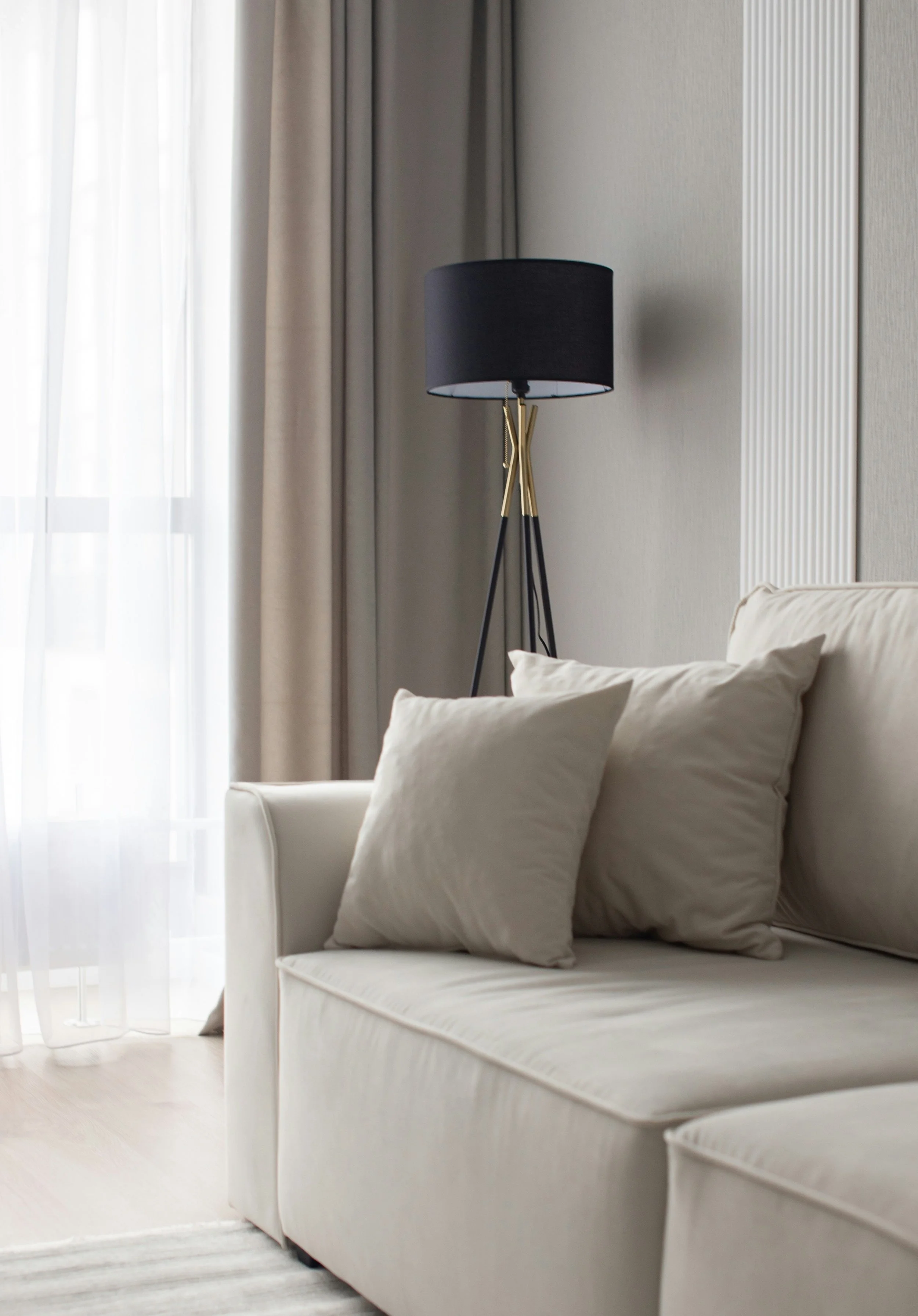 Living room with beige sofa, light beige pillows, a black and gold standing lamp, and beige curtains next to a sheer white curtain.