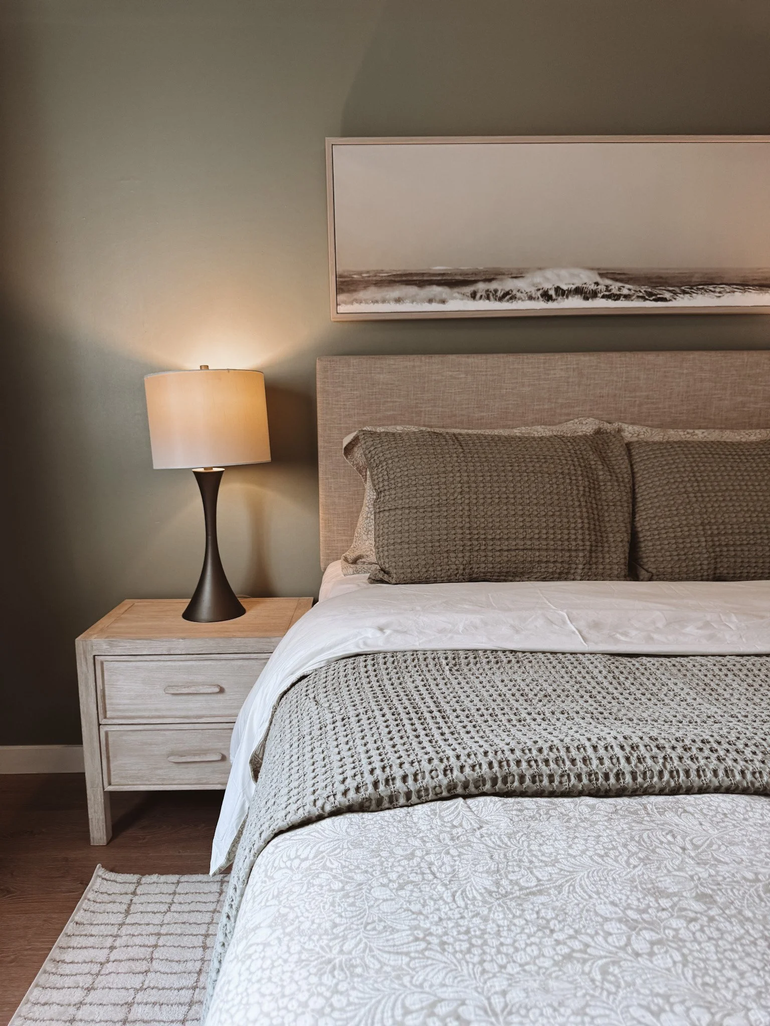 Close-up of a neatly made bed with a beige upholstered headboard, decorative pillows, a white patterned bedspread, and a textured throw blanket. To the left, a wooden nightstand with two drawers and a modern black lamp with a beige lampshade. A framed landscape art piece hanging above the headboard. Greenish wall behind the bed.