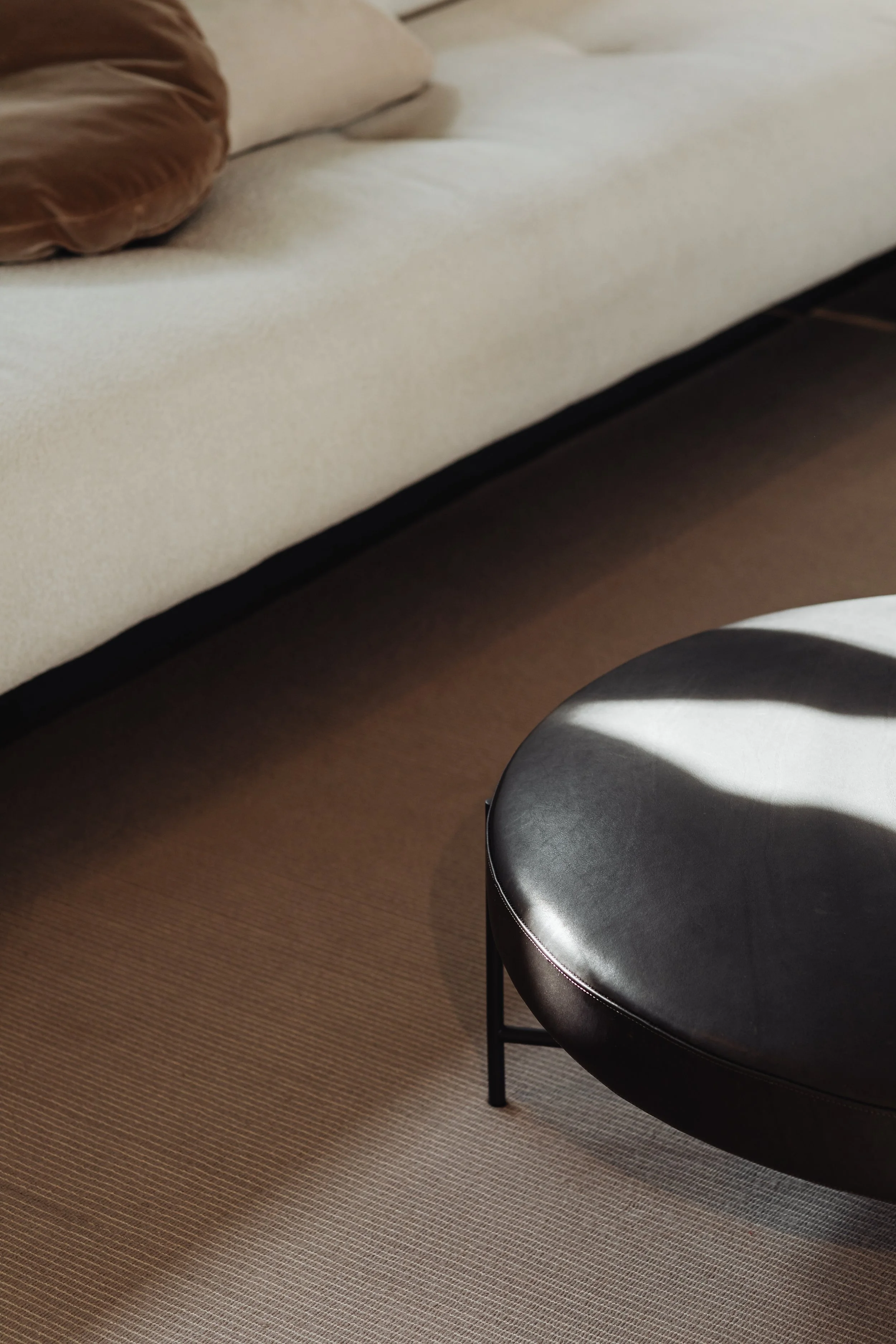 Close-up of a black leather round ottoman and part of a beige sofa with a brown throw pillow on a beige carpeted floor.