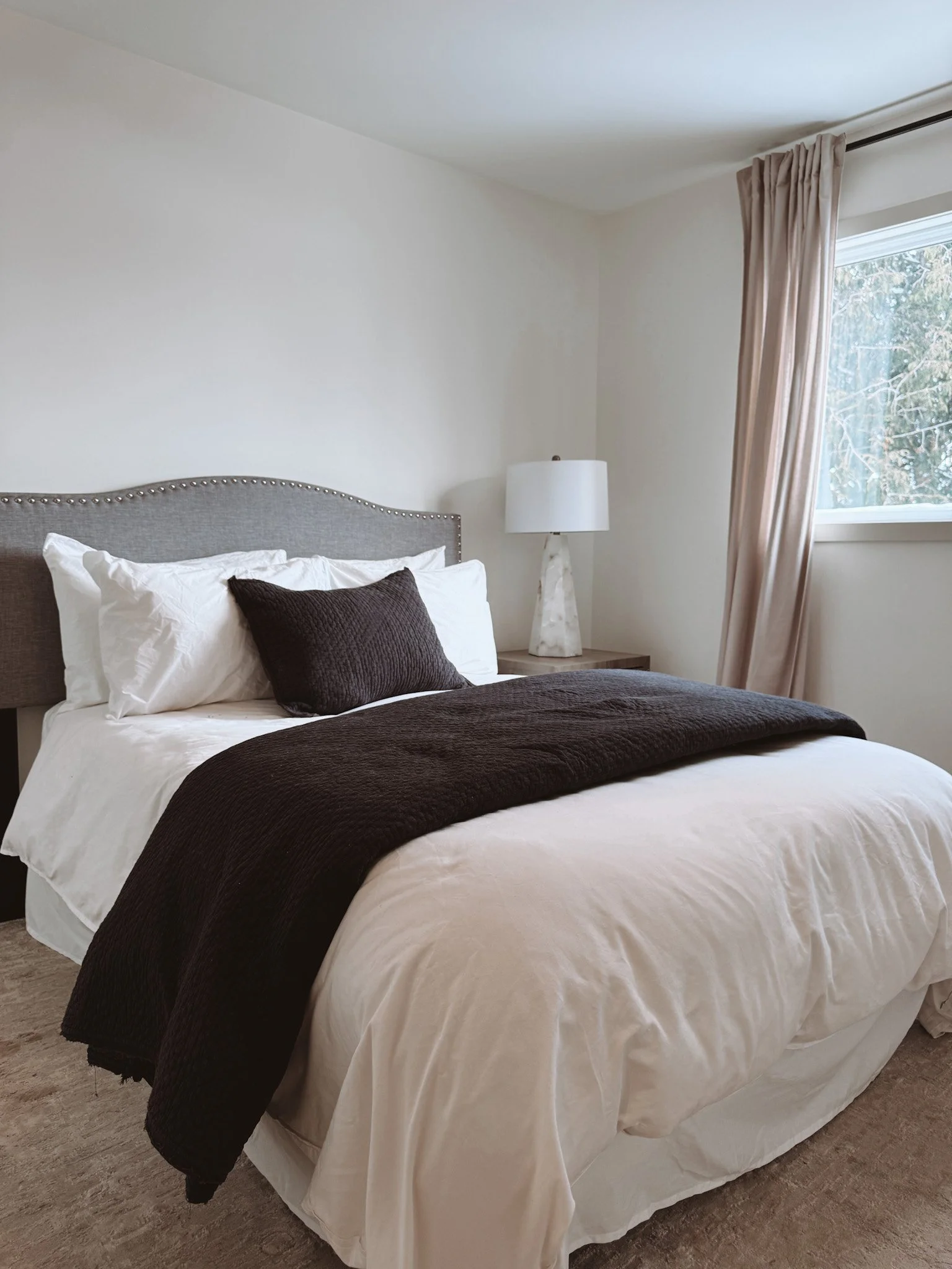 A neatly made bed with white sheets, a black blanket, and a black pillow, in a bedroom with a window, beige curtains, a white lamp on a wooden nightstand, and a view of trees outside.