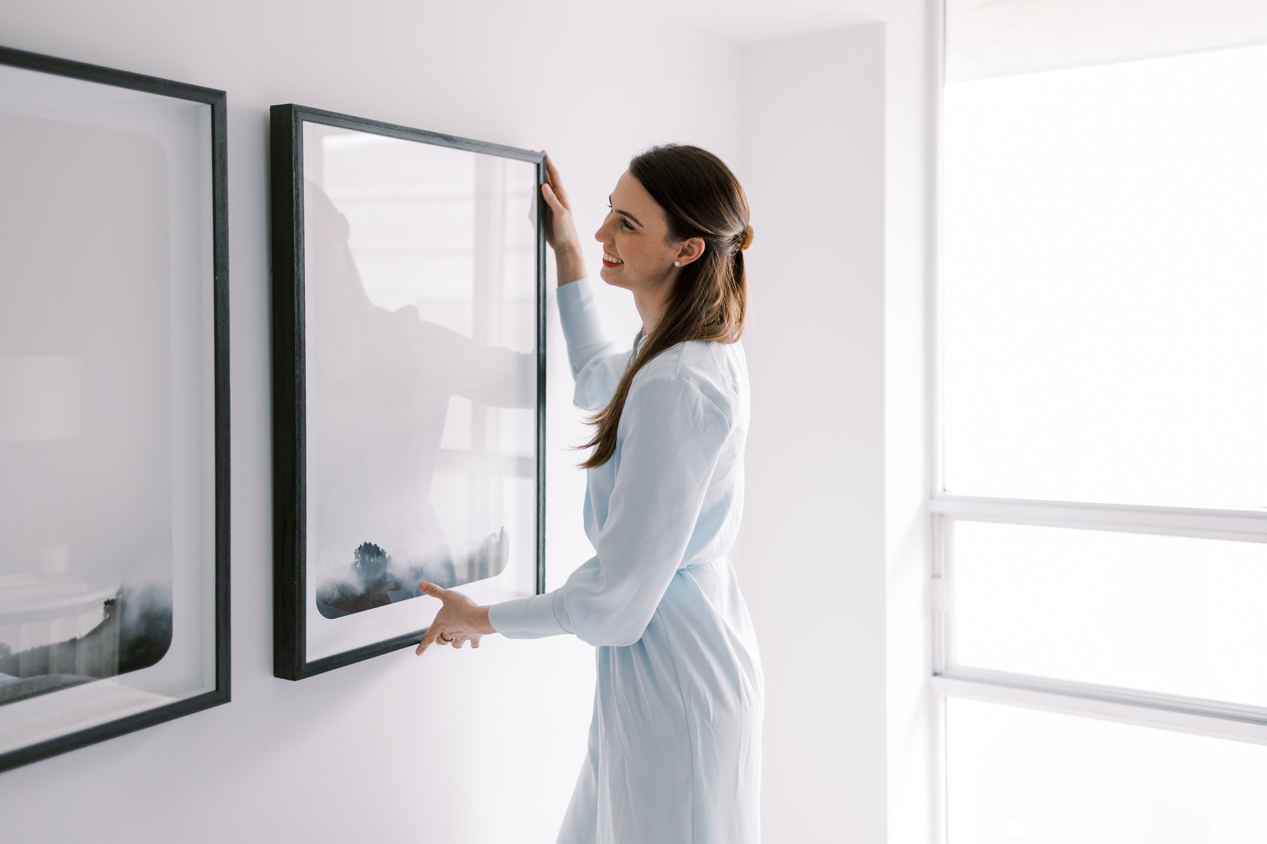 A woman in a white dress hanging paintings on a white wall in a bright room.