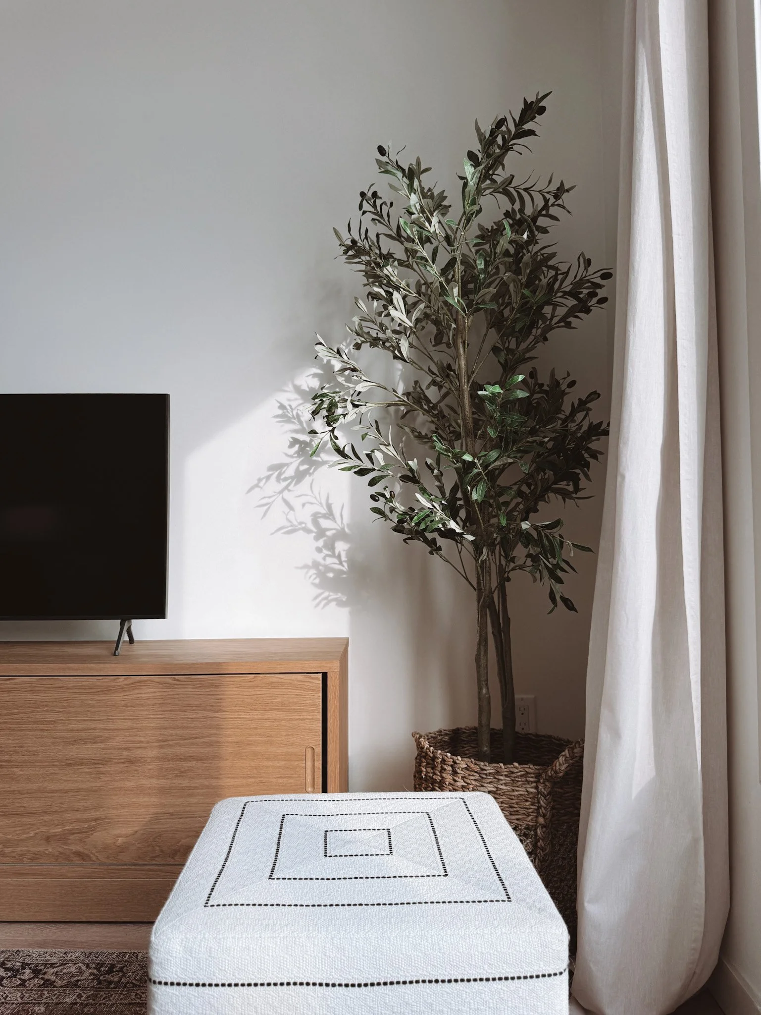 A living room corner with a wooden TV stand, a black flat-screen TV, a tall potted plant with dark green leaves, a woven basket, a white curtain, and a white ottoman with black stitched square patterns.
