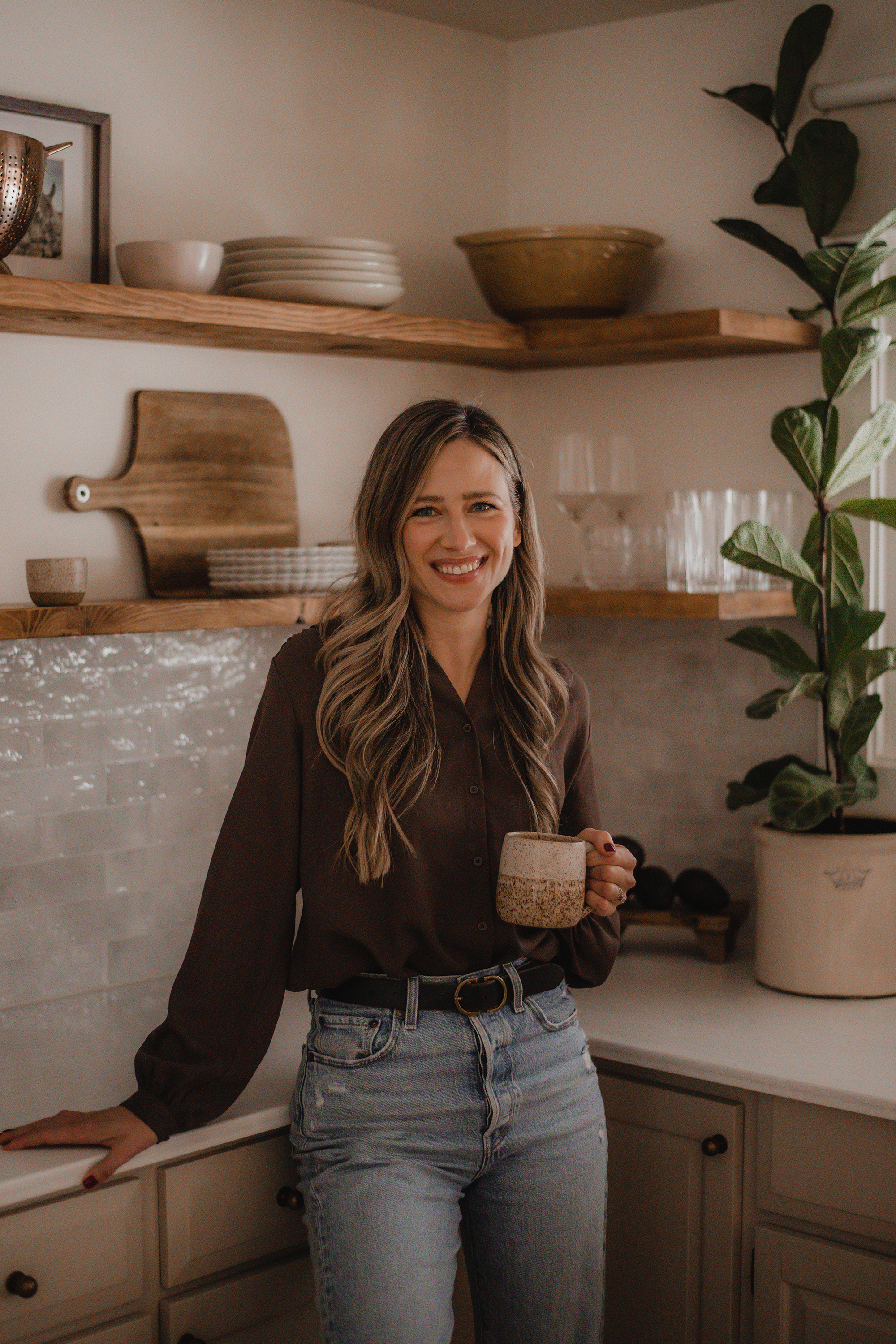 A woman with long wavy hair smiling and holding a ceramic mug in a cozy kitchen with wooden shelves, bowls, plates, a potted plant, and neutral-colored decor.