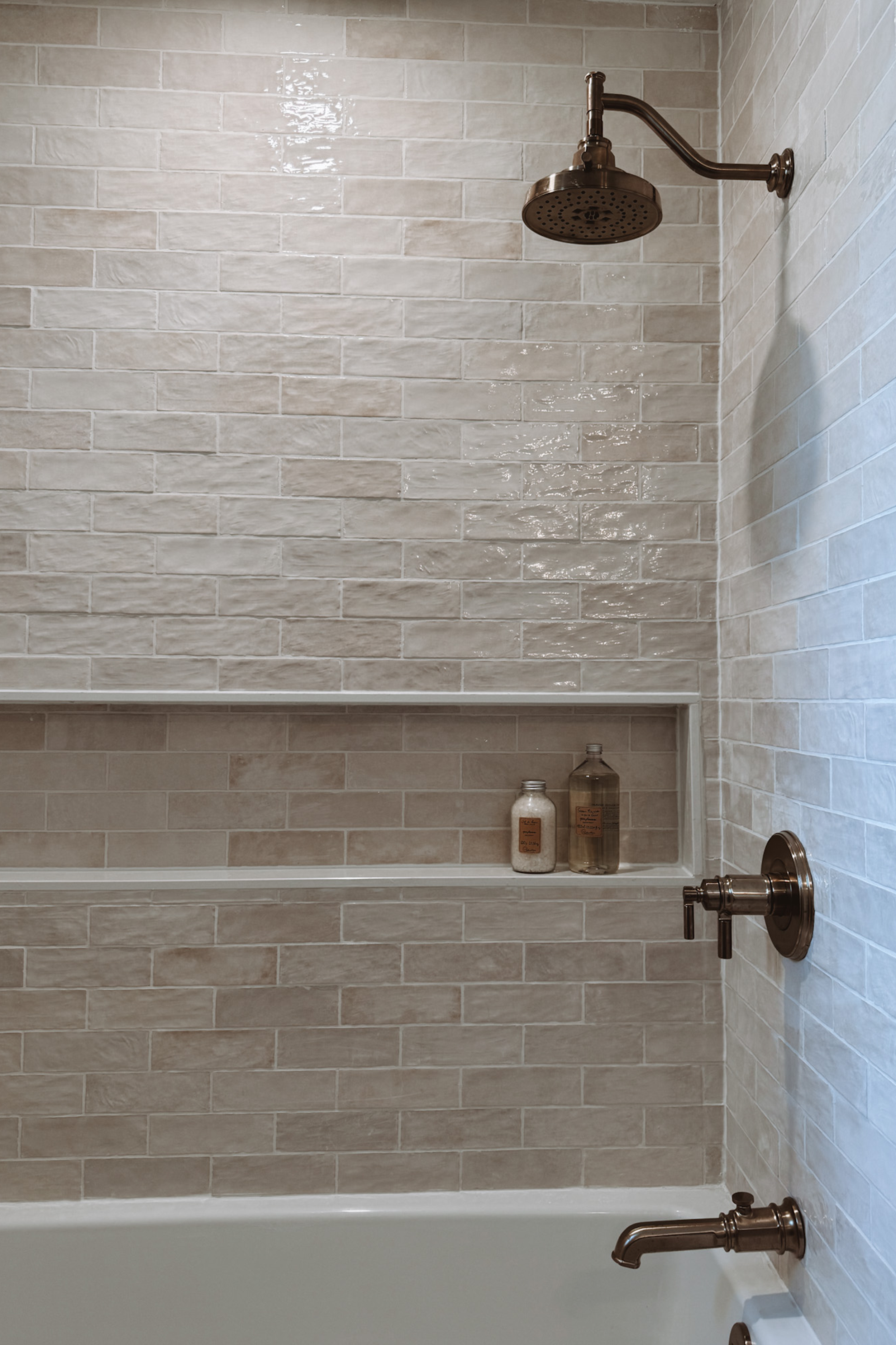 A bathroom shower with beige tiled walls, a metal showerhead, and built-in shelf with bottles.