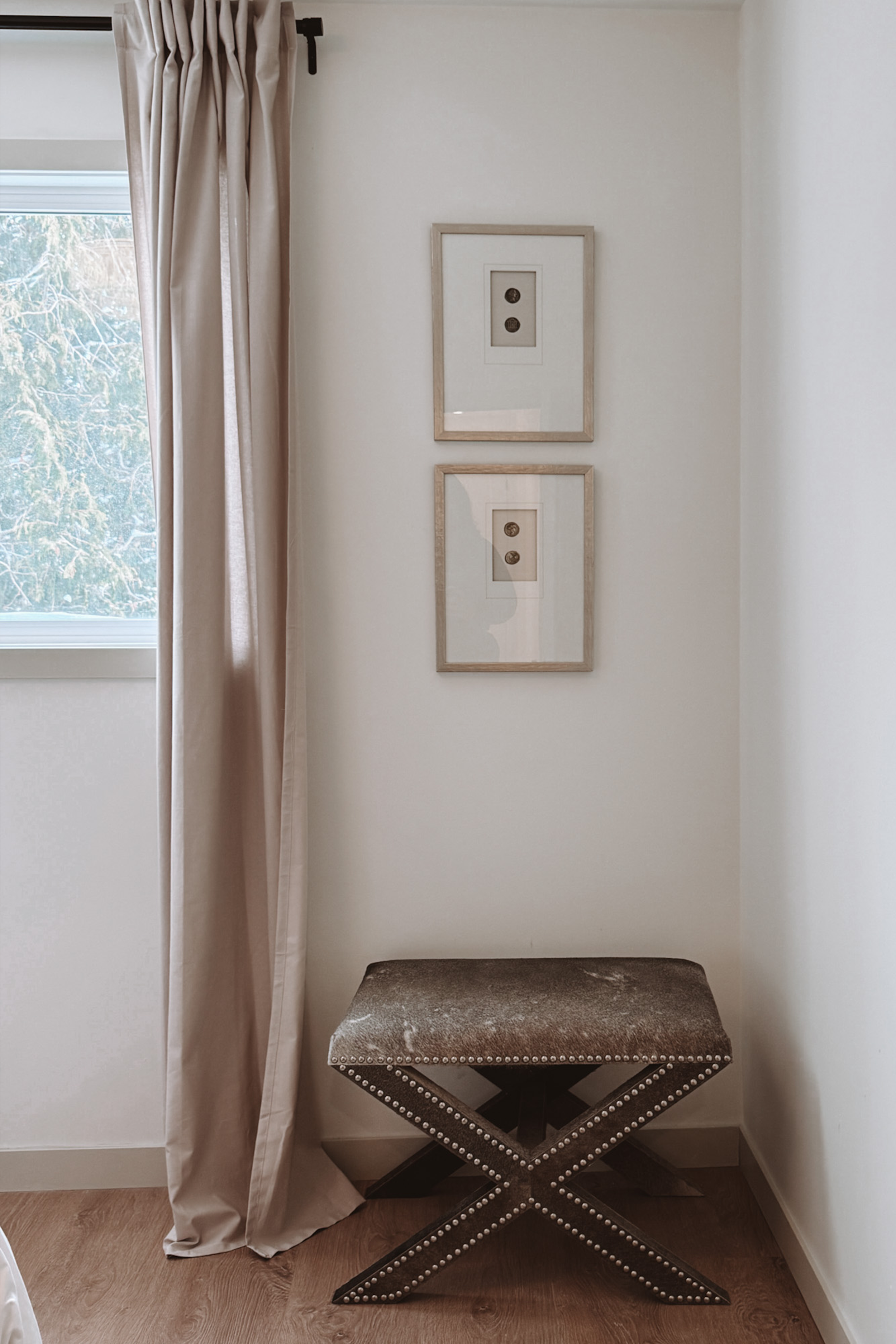 A corner of a room with cream curtains partially covering a window, two framed pictures with buttons on the wall, and a brown upholstered bench with nailhead trim on the seat and crossed legs.