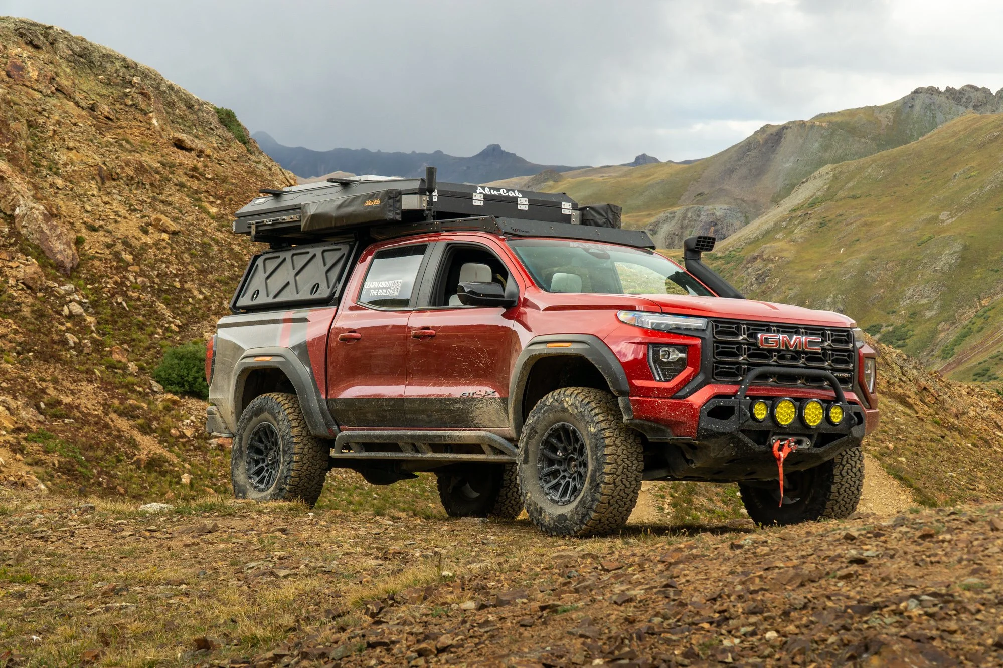 Red GMC off-road vehicle with large tires and mounted equipment driving in a mountainous terrain with rocky ground and green hills under cloudy sky.