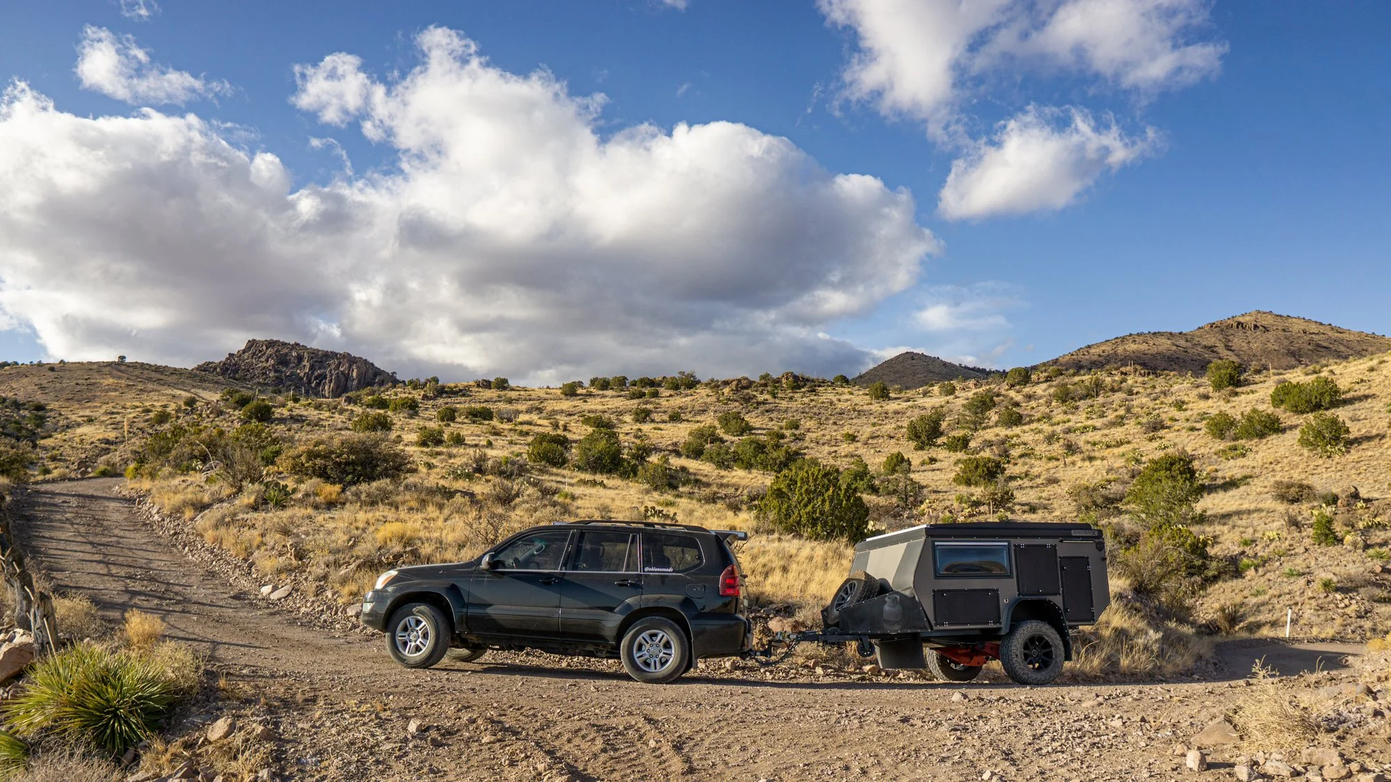 A black SUV towing a compact black off-road camper trailer on a dirt trail in a semi-arid landscape with small bushes, distant hills, and a partly cloudy sky.