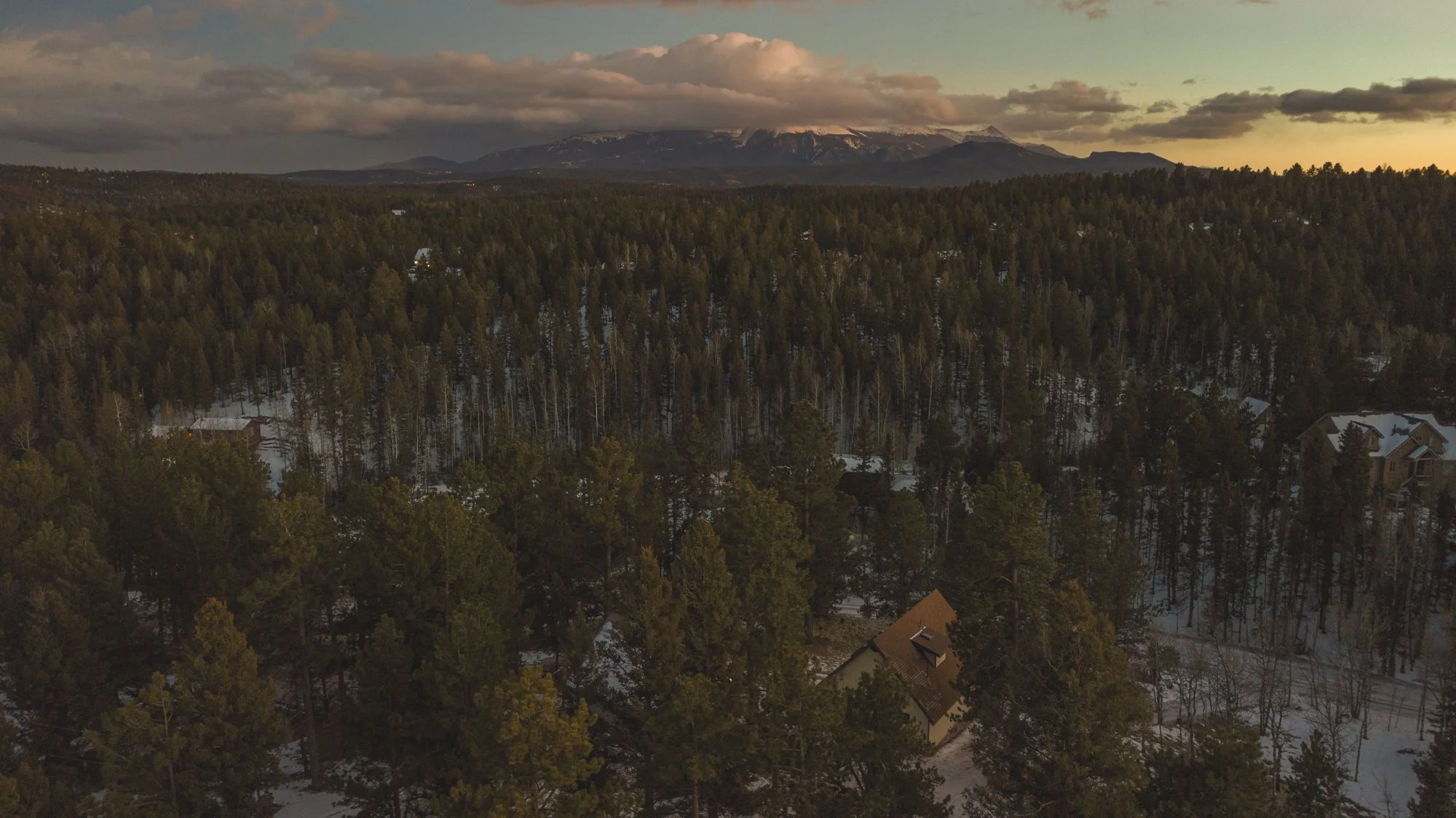 Aerial view of a forested landscape with snow patches, residential houses, distant mountains, and a cloudy sky at sunset.