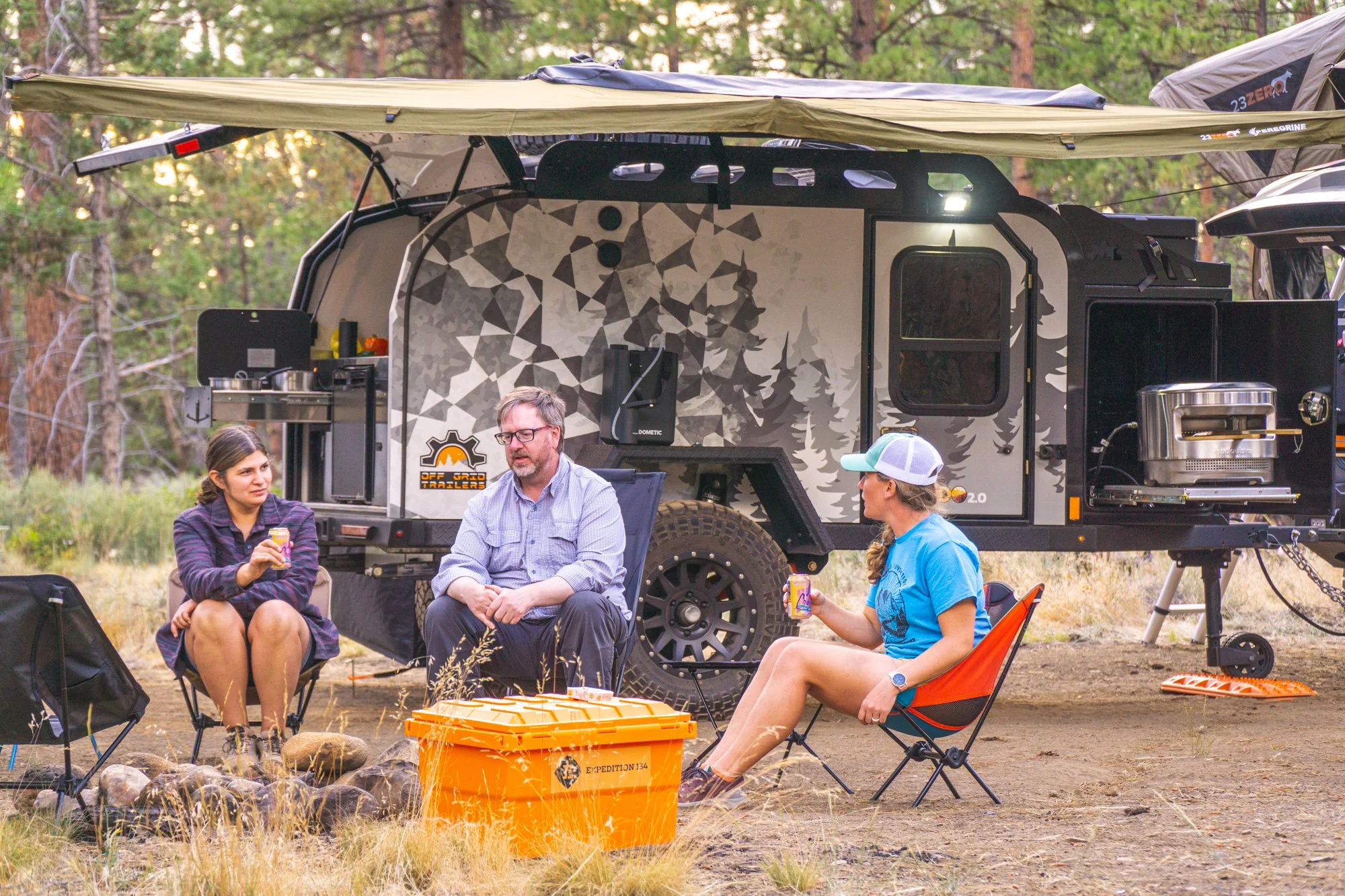 Three people sitting in camp chairs around a campfire near a trailer in a forested area, enjoying drinks.