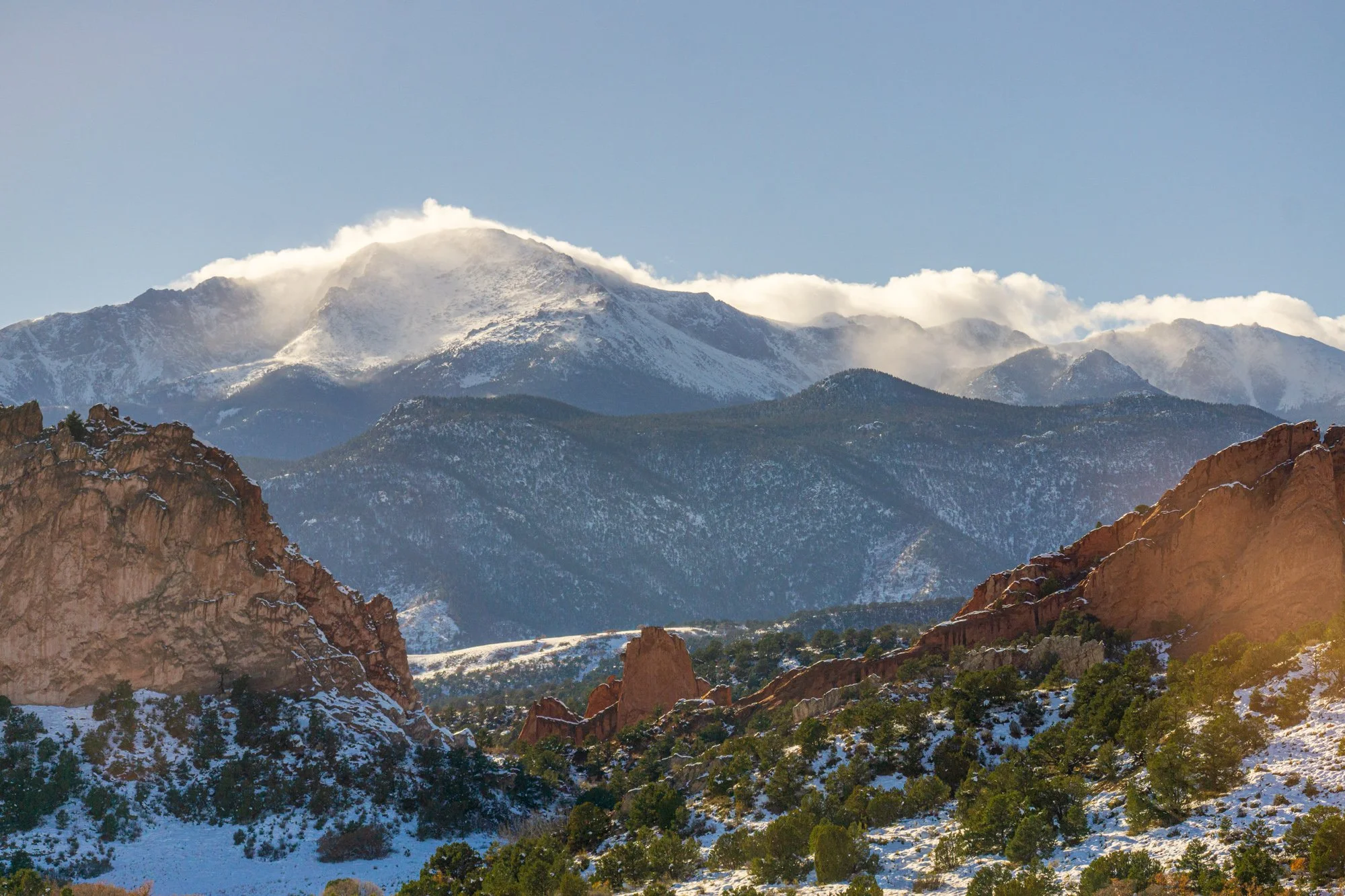 Snow-capped mountains with rocky formations and green trees in a sunny landscape.