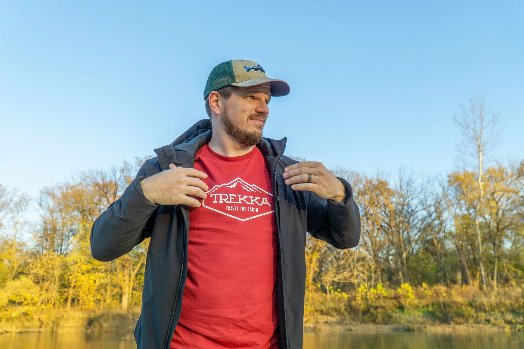 A man outdoors near a body of water with trees showing fall foliage, wearing a red T-shirt and a black jacket, adjusting his jacket, under a clear blue sky.