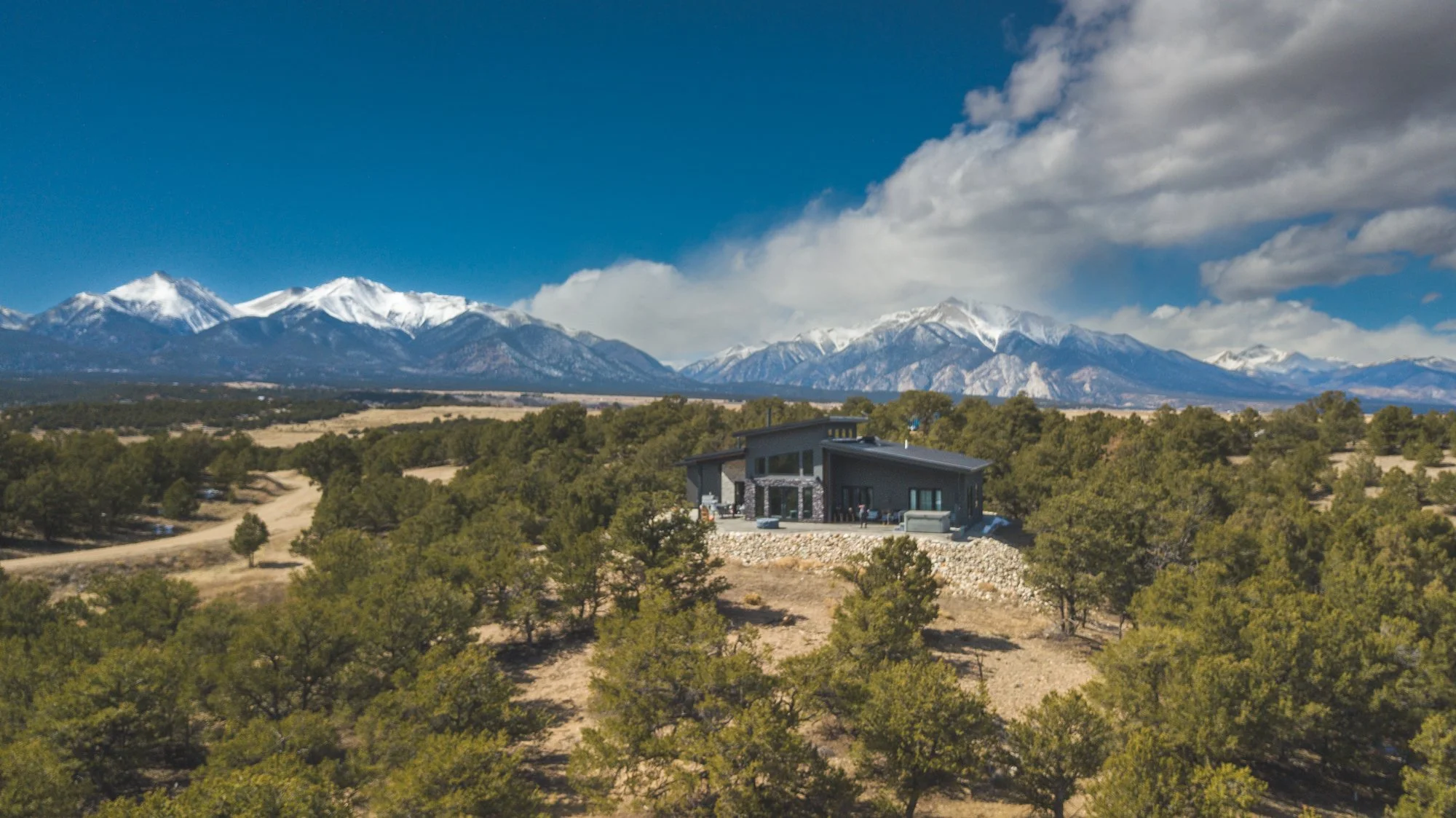 Modern house on a hillside surrounded by trees with snow-capped mountains in the background.