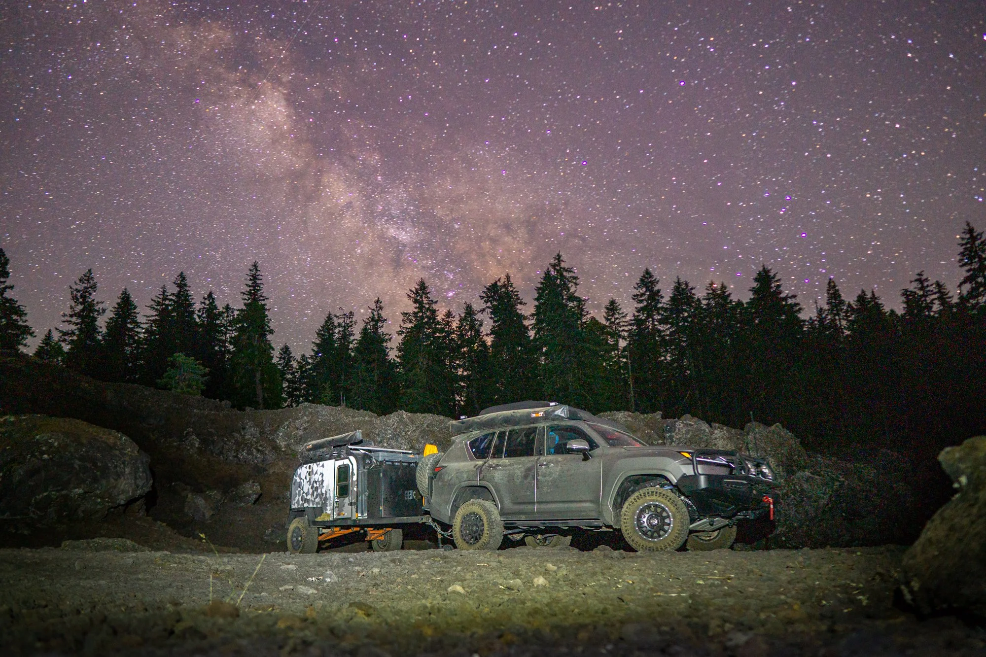 Off-road vehicle and small trailer parked on rocky ground beneath a starry night sky with visible Milky Way galaxy and a forest of tall trees in the background.