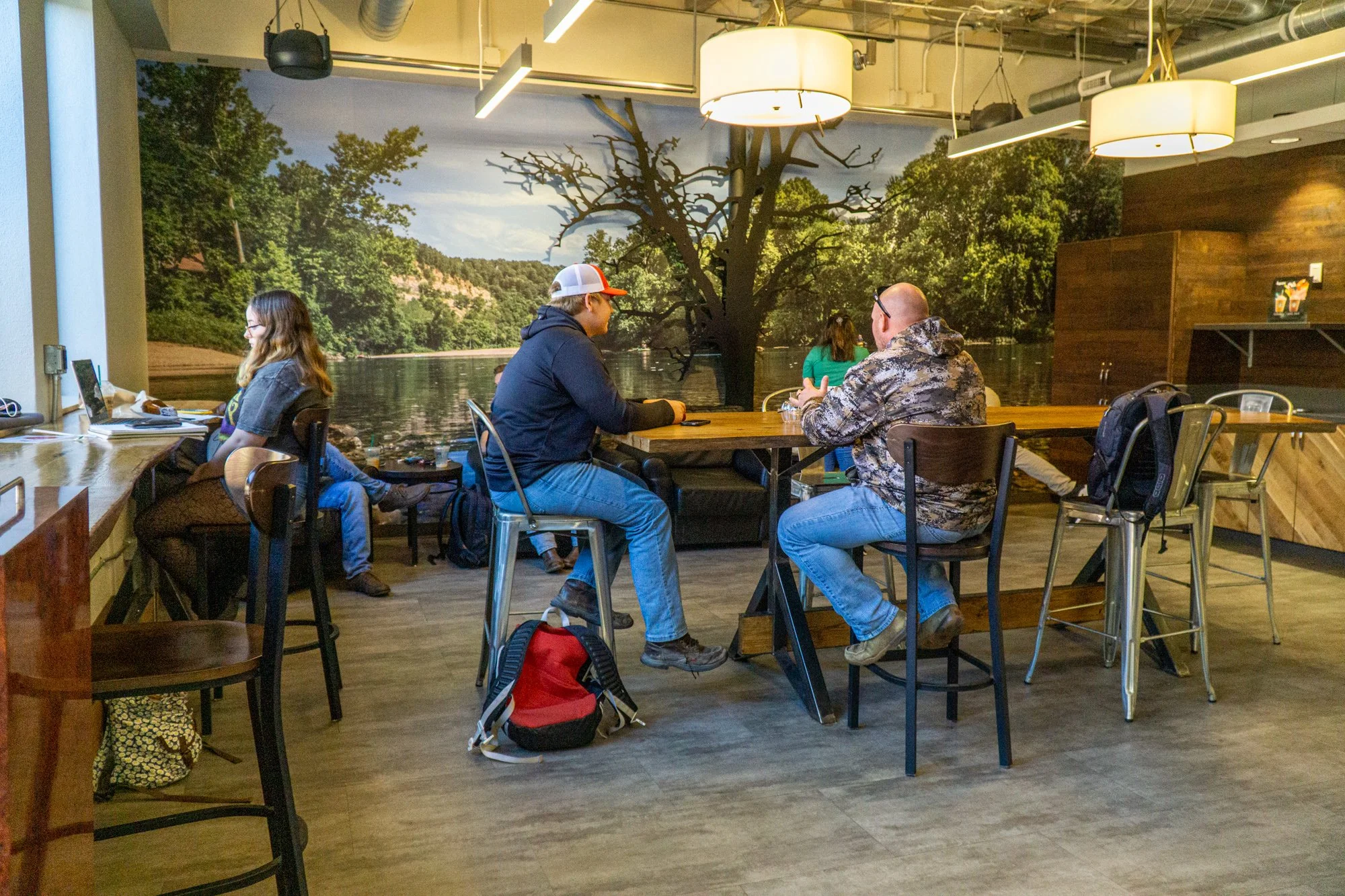 People sitting at a table in a cafe with a large wall mural of a river, trees, and a leafless tree in the background.