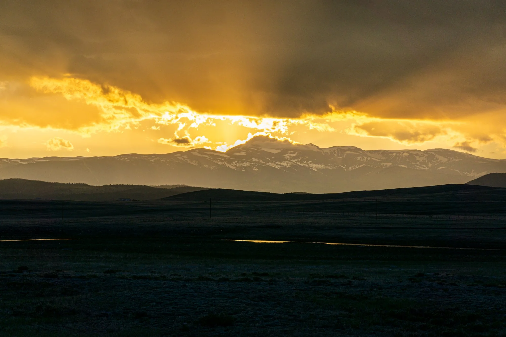 A landscape at sunset showing dark rolling hills in the foreground and a mountain range with snow patches in the background, with golden sunlight breaking through dark clouds in the sky.