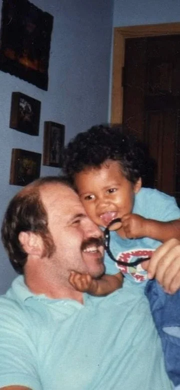 A man with curly hair and a beard holding a young child with curly black hair, both smiling and playing with a pair of glasses indoors.
