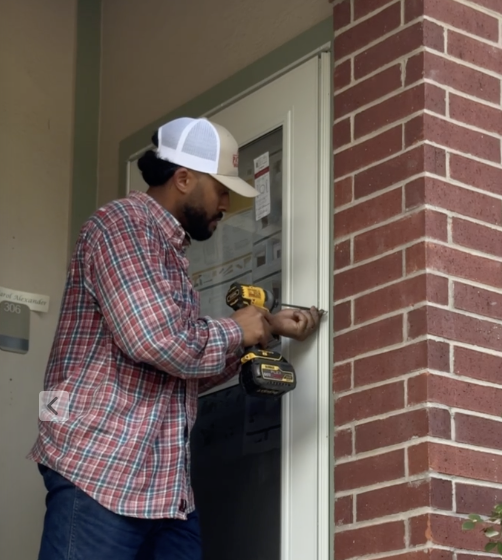 A man wearing a plaid shirt, baseball cap, and glasses installing a doorbell or intercom on a white door next to a brick wall using a power drill.