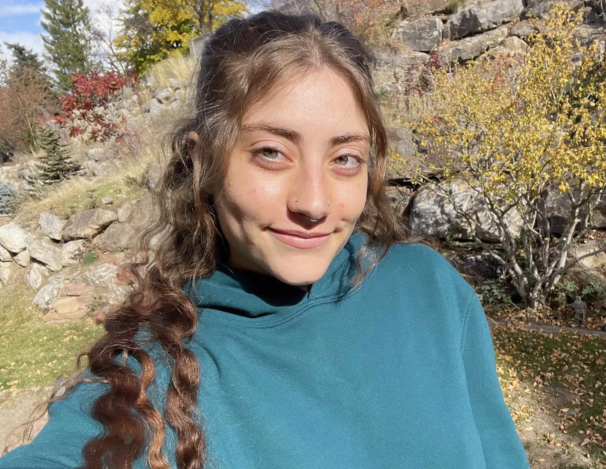 A young woman (Lindsey) outdoors in a sunny setting with trees and rocks in the background, wearing a teal hoodie and smiling at the camera.